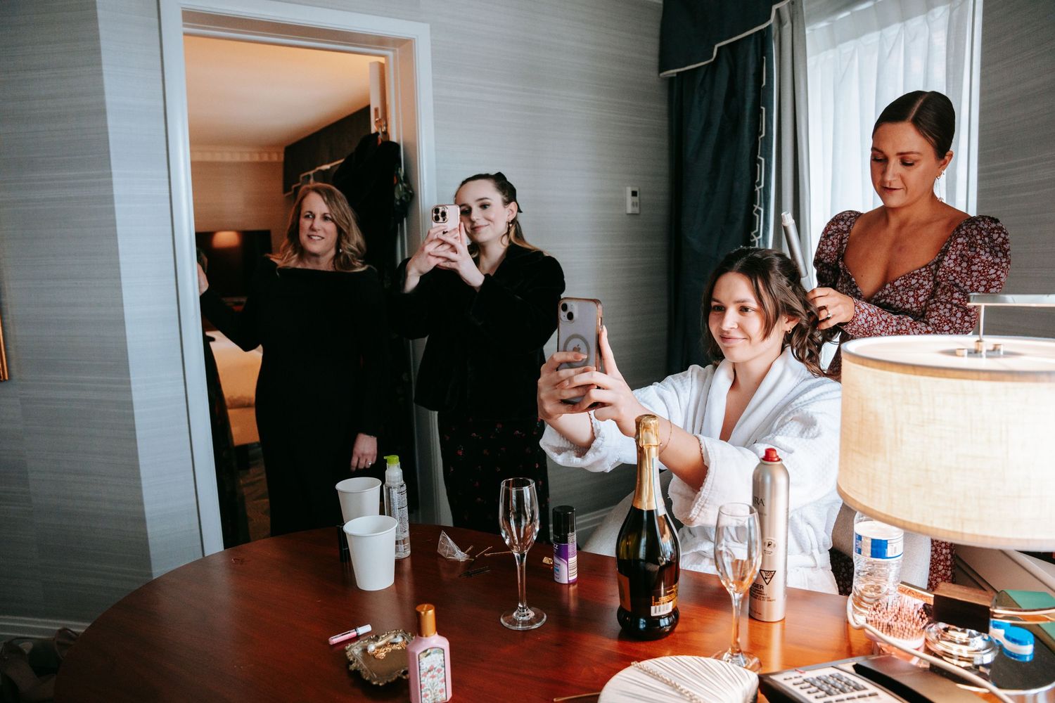 Bridesmaids taking selfies and getting ready together in a hotel room with champagne on the table.
