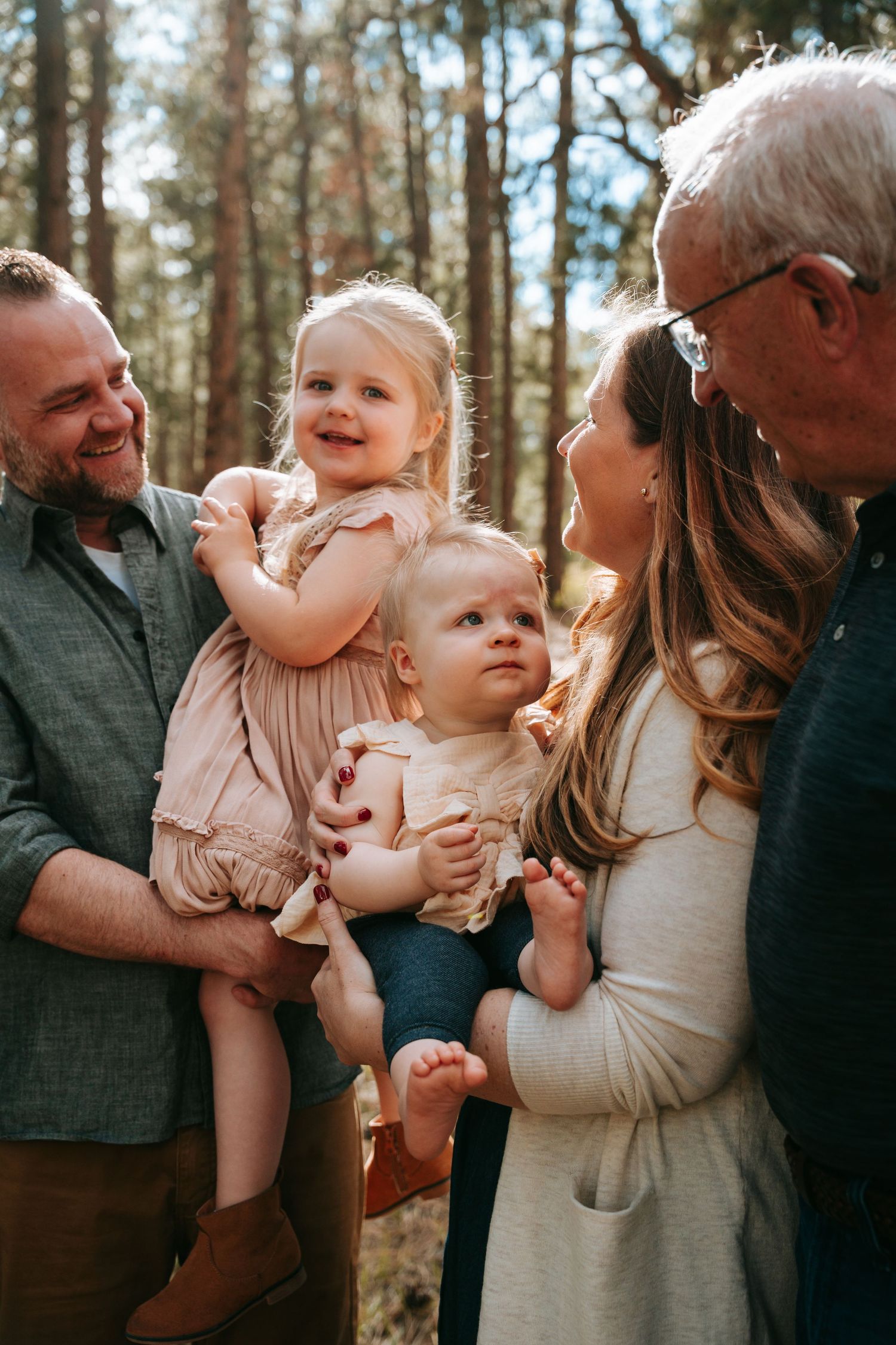 Family with two toddlers smiling outdoors in a sunlit pine forest during a warm family photo session.