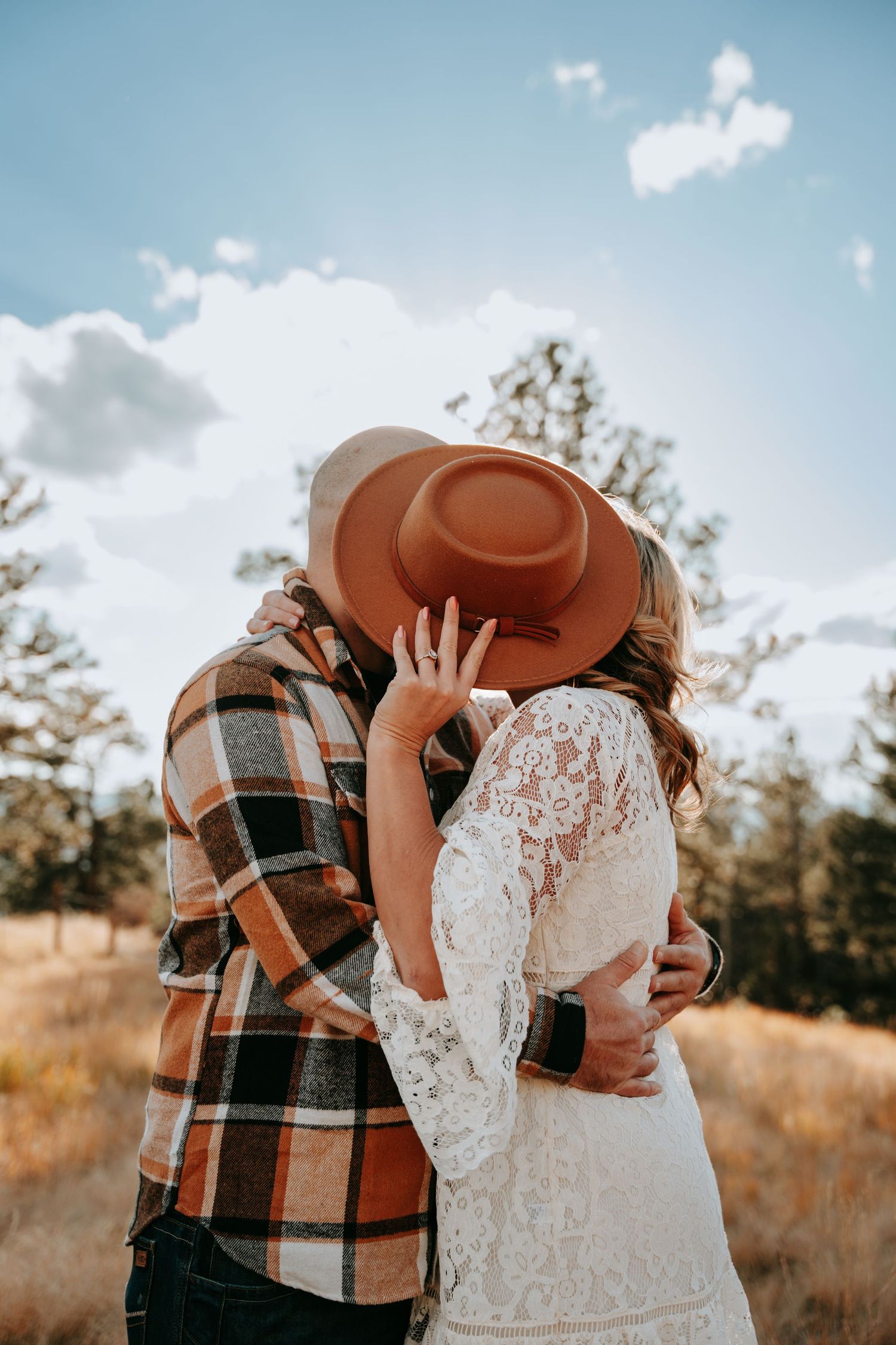 Couple sharing a kiss hidden behind brown wide-brim hat in sunny outdoor field.
