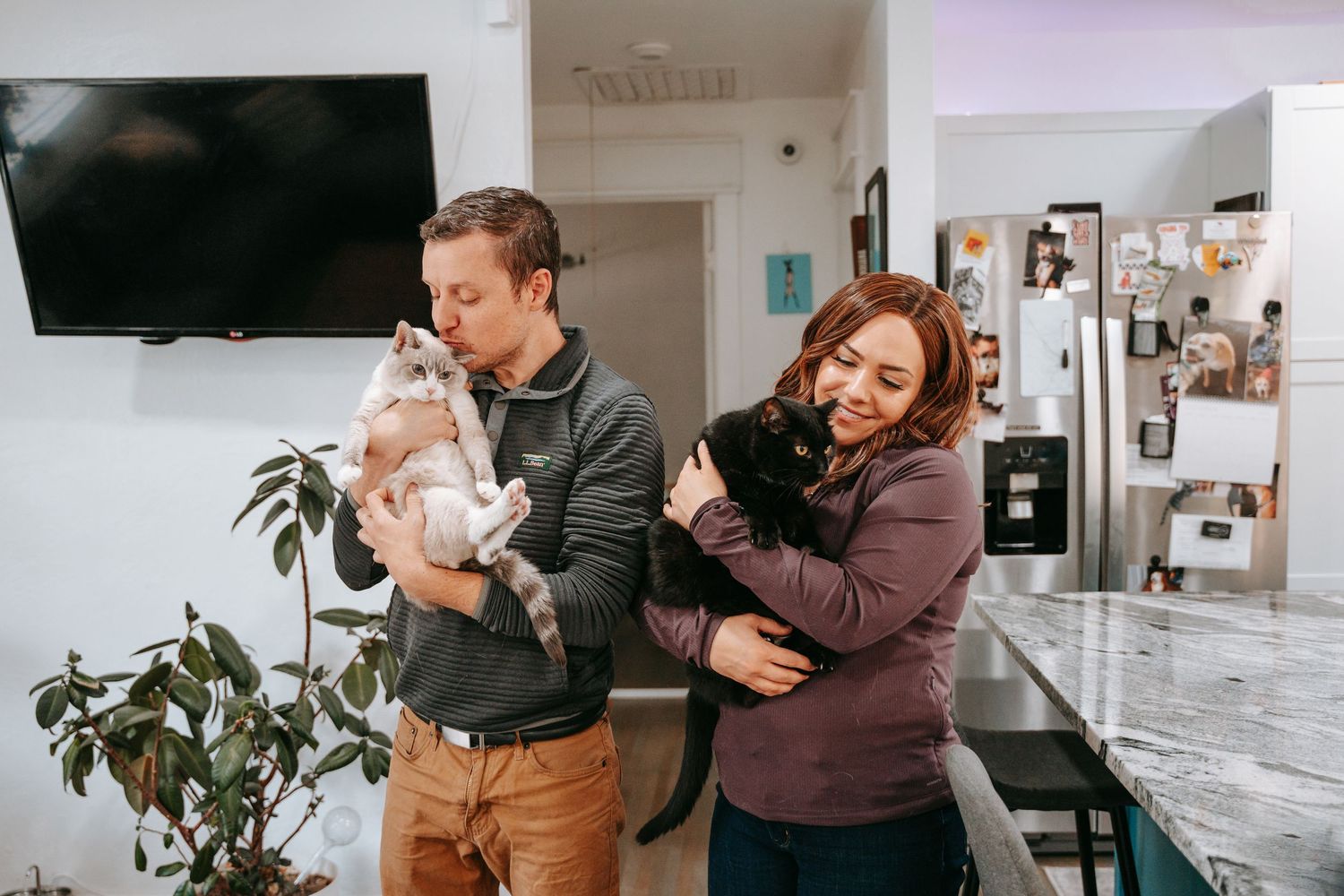 A happy couple stands in their kitchen, each holding a cat, smiling and enjoying their pets.