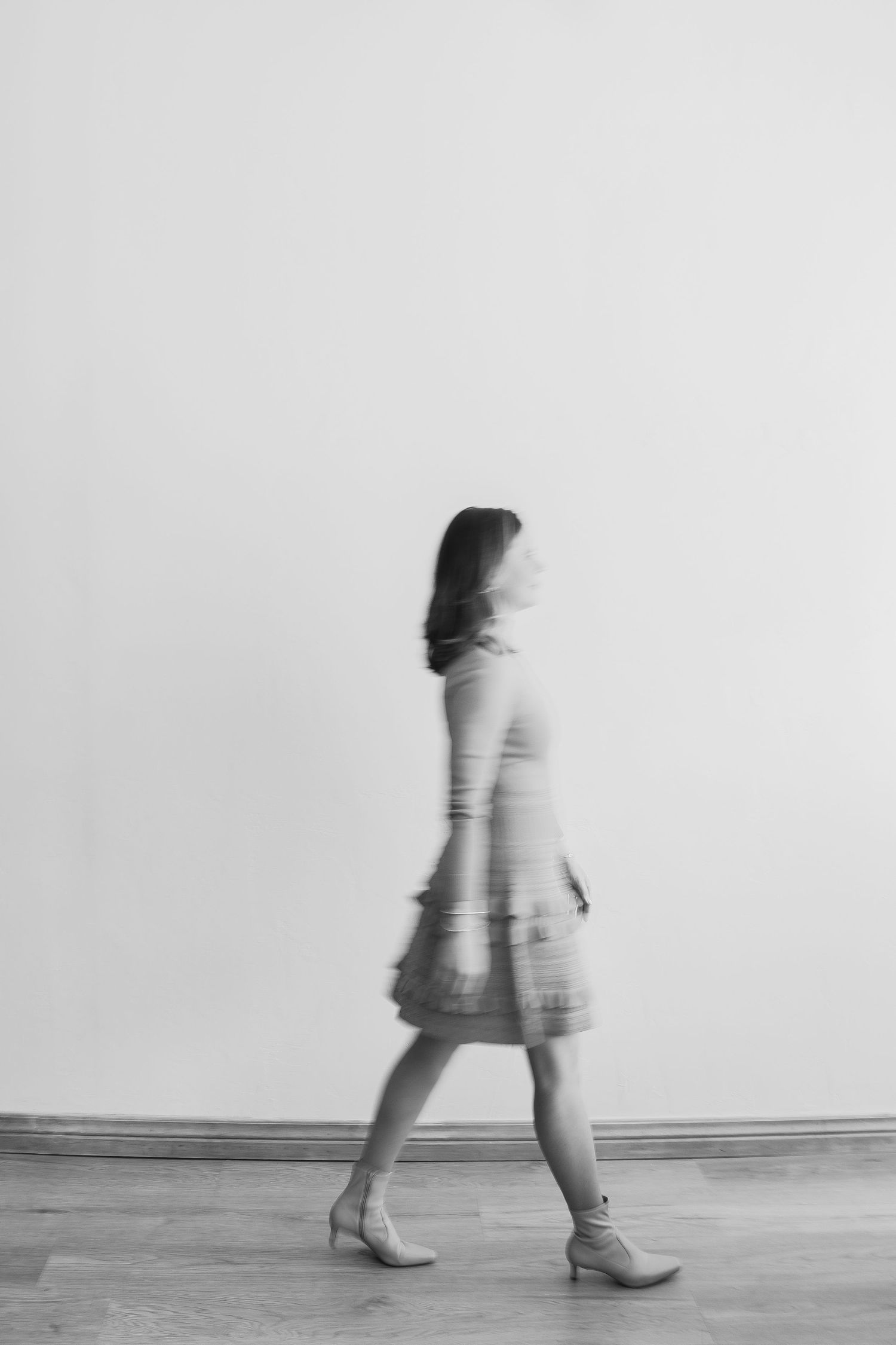 Black and white motion blur artistic photo of woman walking in tiered dress and heels against white wall.