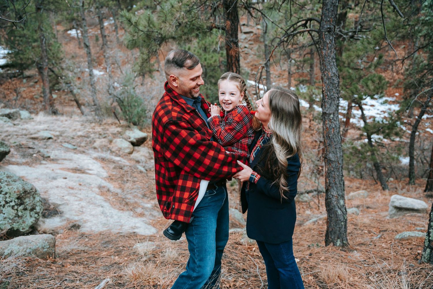 Happy family of three in red plaid outfits laughing together among snow-dusted forest trees.