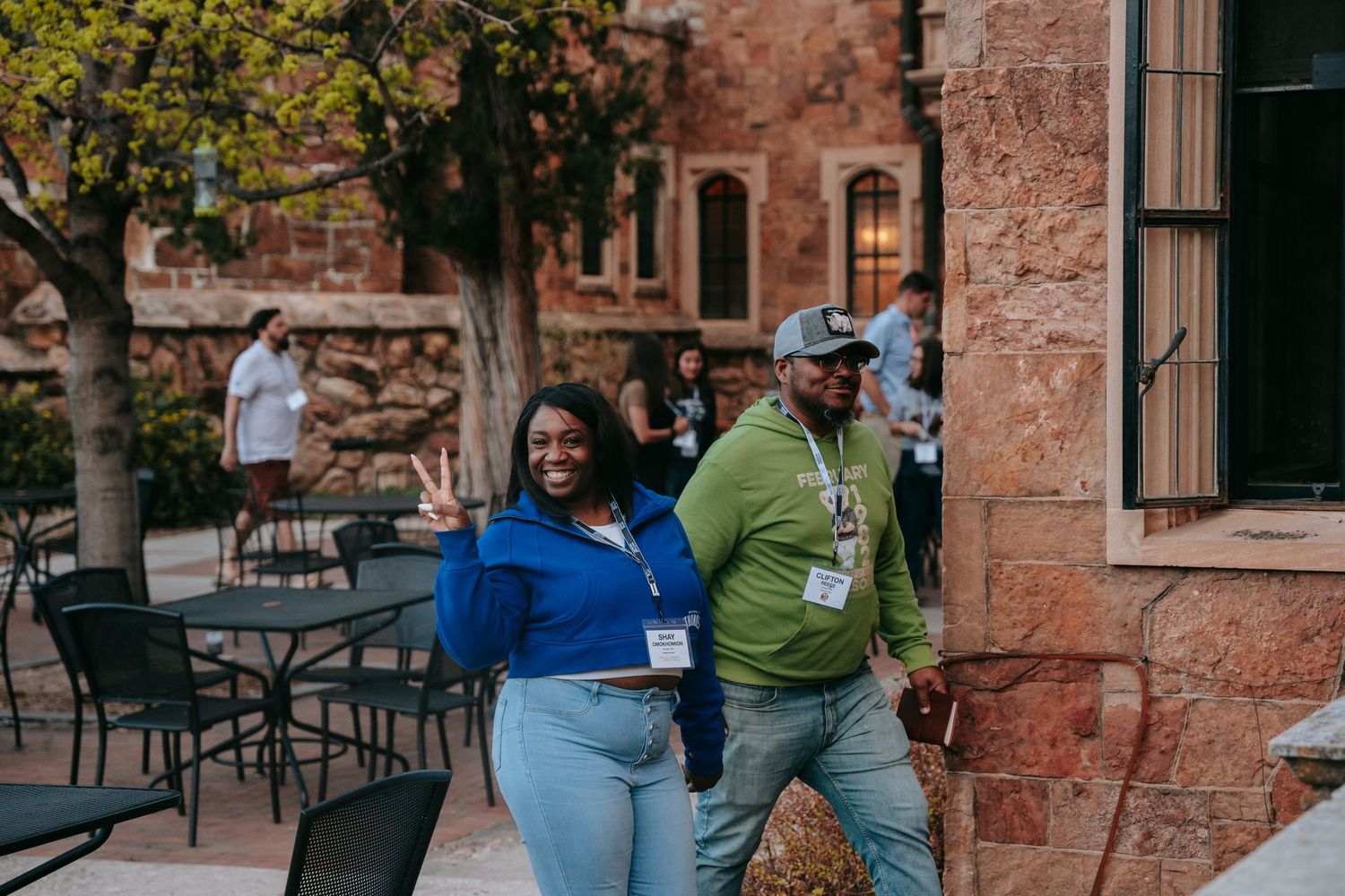 Two smiling people wearing lanyards walk outside a historic stone building at an outdoor event.