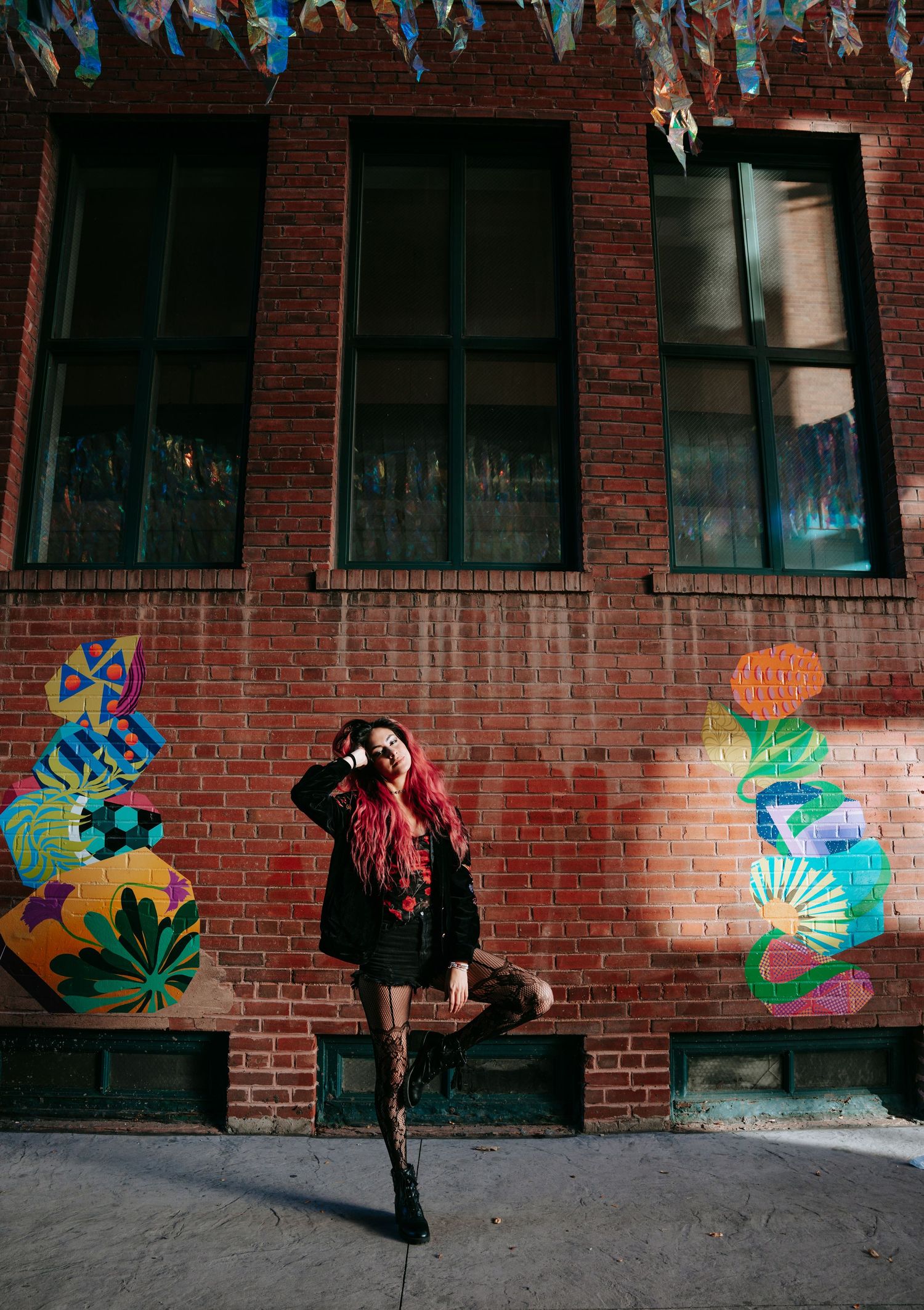 Woman with red hair posing confidently in front of colorful murals on a brick building wall.