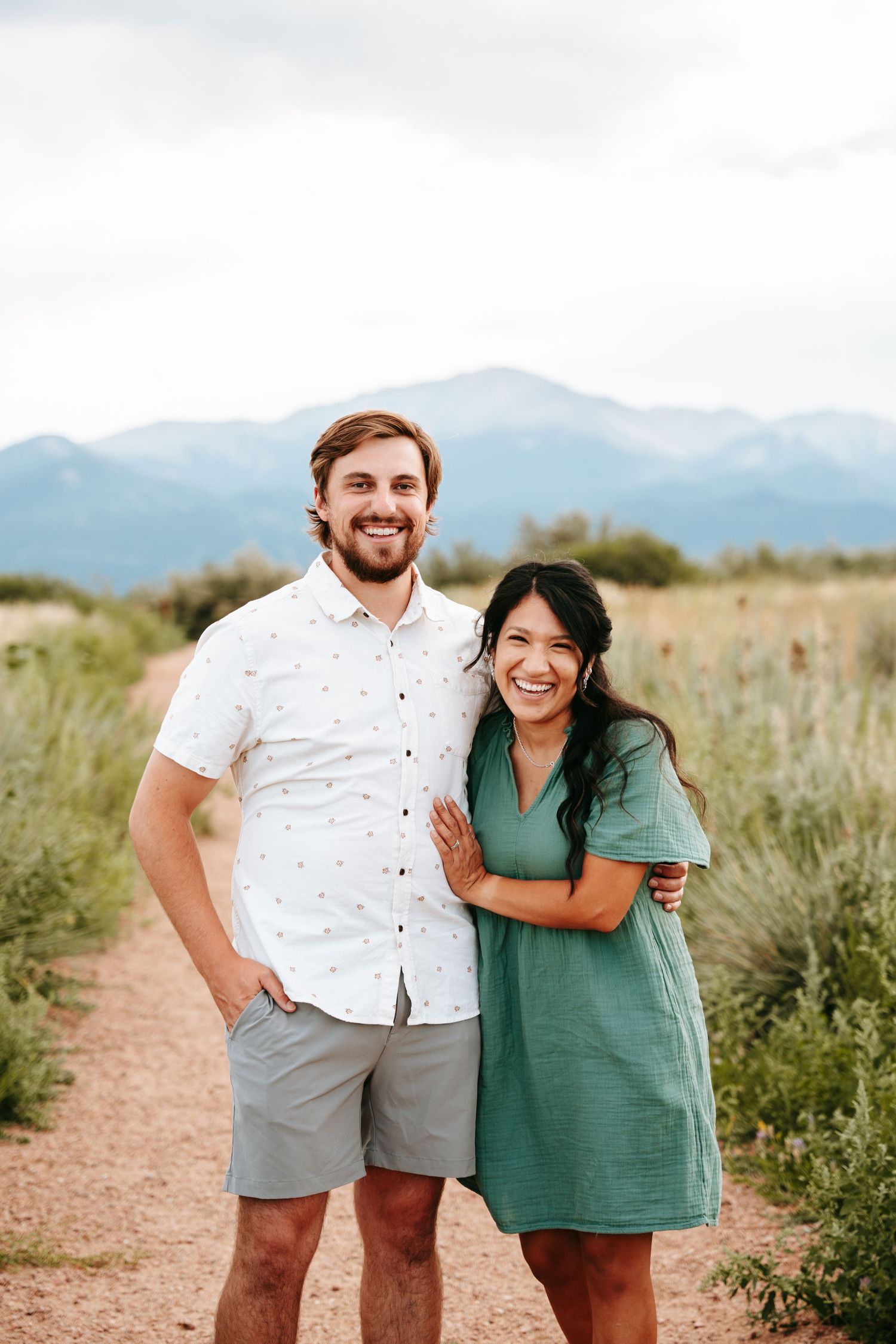 A joyful couple poses together on a dirt path with stunning mountain views during an outdoor engagement session.