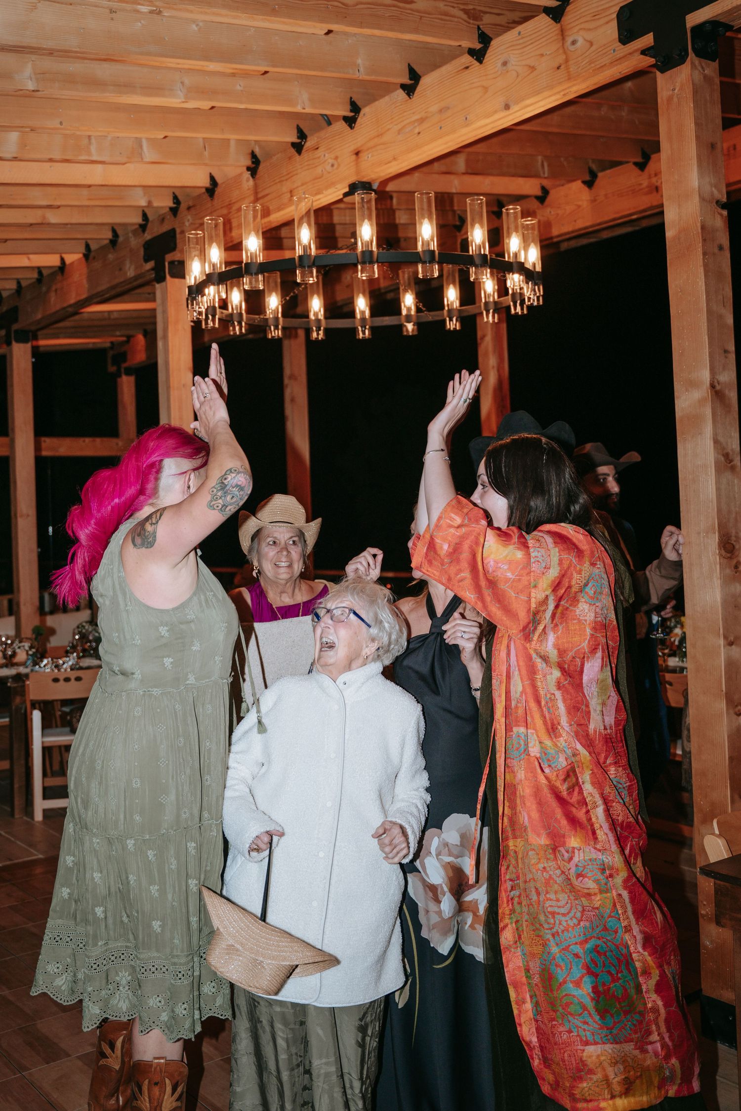 Wedding guests dancing and raising hands under a rustic chandelier in a wooden barn venue.