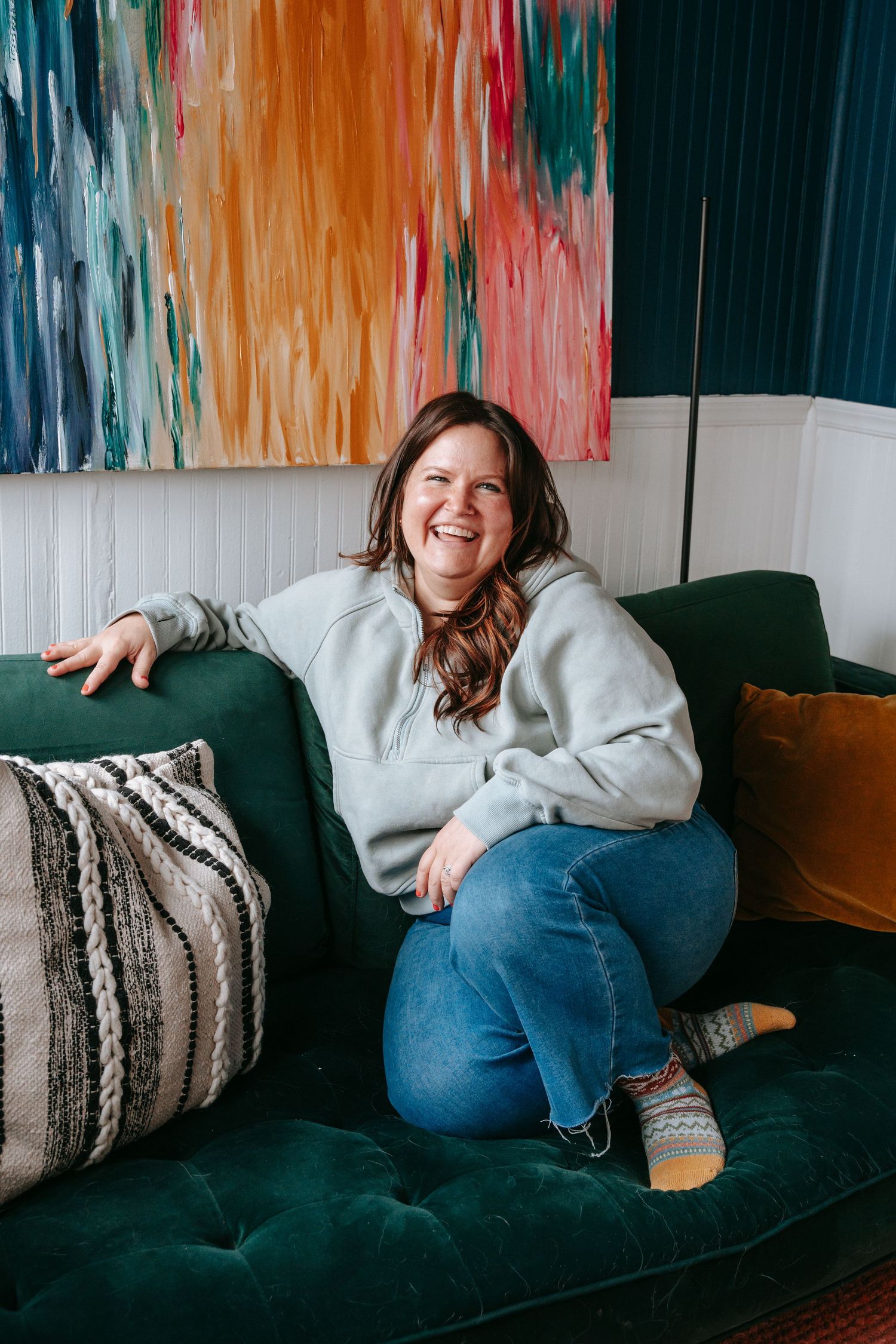 Smiling woman sitting on a green velvet sofa with colorful abstract painting on the wall behind her.