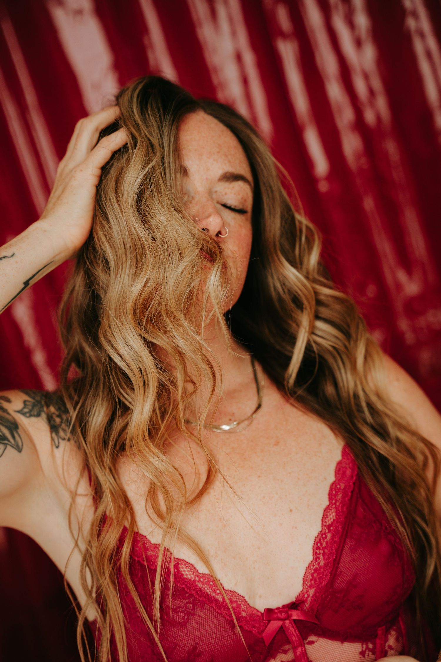 Woman with long wavy blonde hair in red lace lingerie poses against a deep red backdrop with eyes closed.