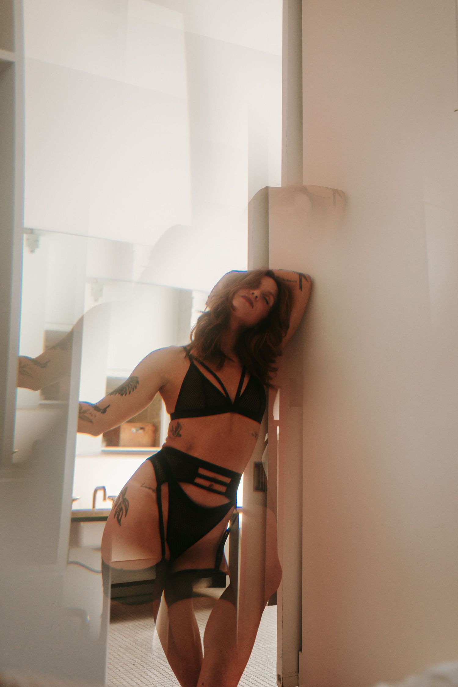 Woman in black lingerie with tattoos leaning against a white wall in a bright modern interior.