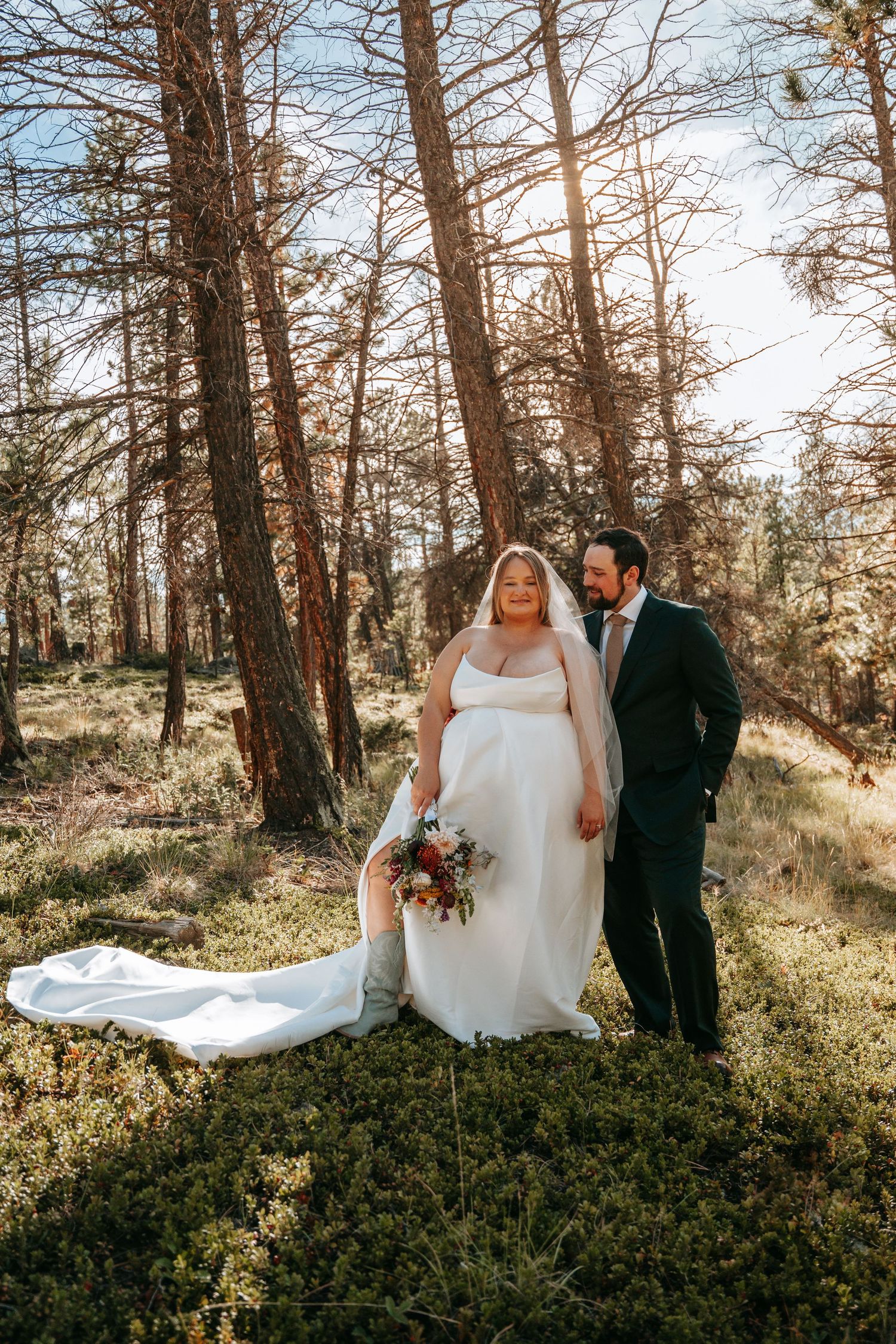 Bride and groom pose in sunlit pine forest, bride in white gown with train holding colorful bouquet.