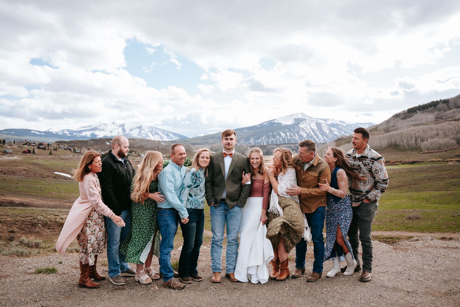 Wedding party group laughing together outdoors with stunning snow-capped mountain landscape behind them.