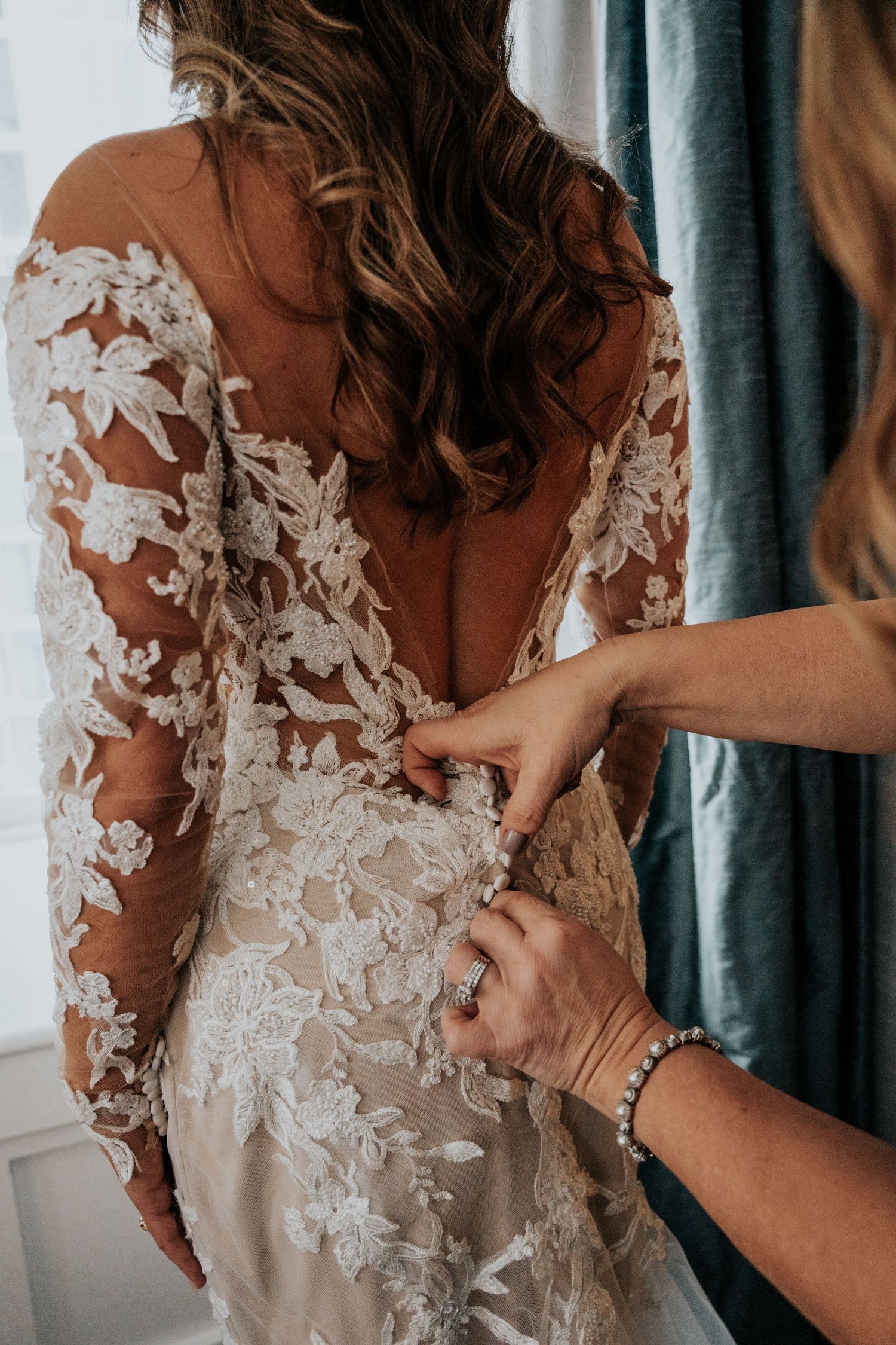 Helper fastening buttons on bride's intricate lace long-sleeve wedding dress from behind.