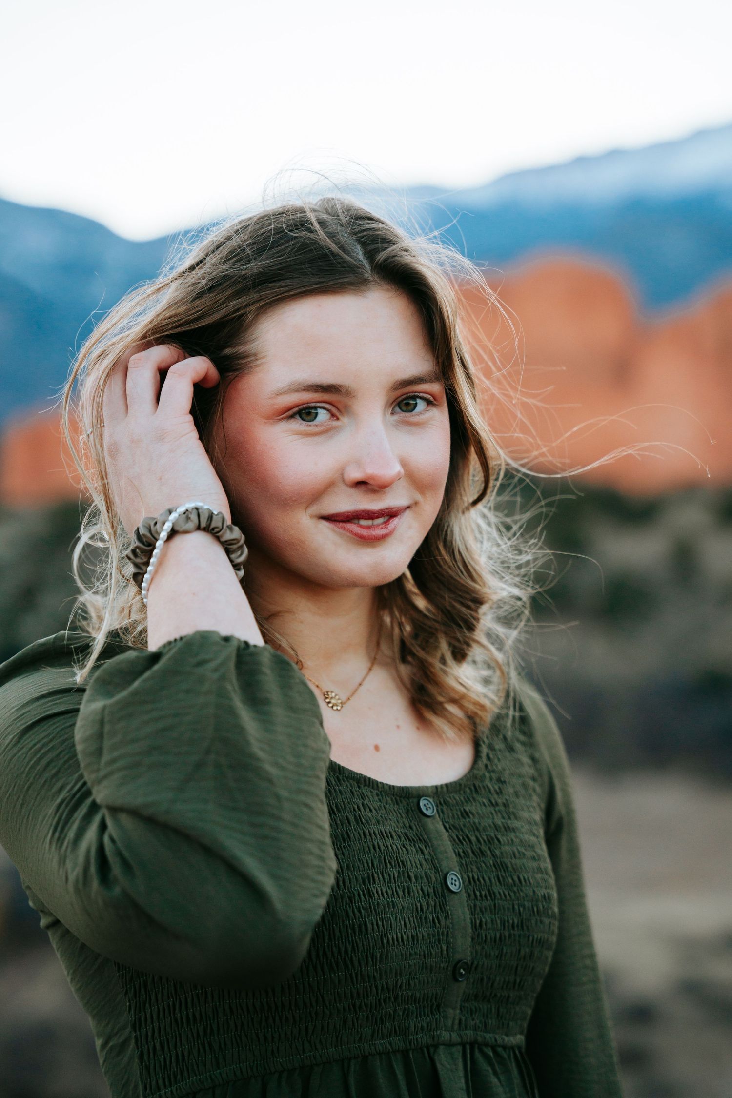 Young woman in green smocked dress touching hair with red rocks in background.
