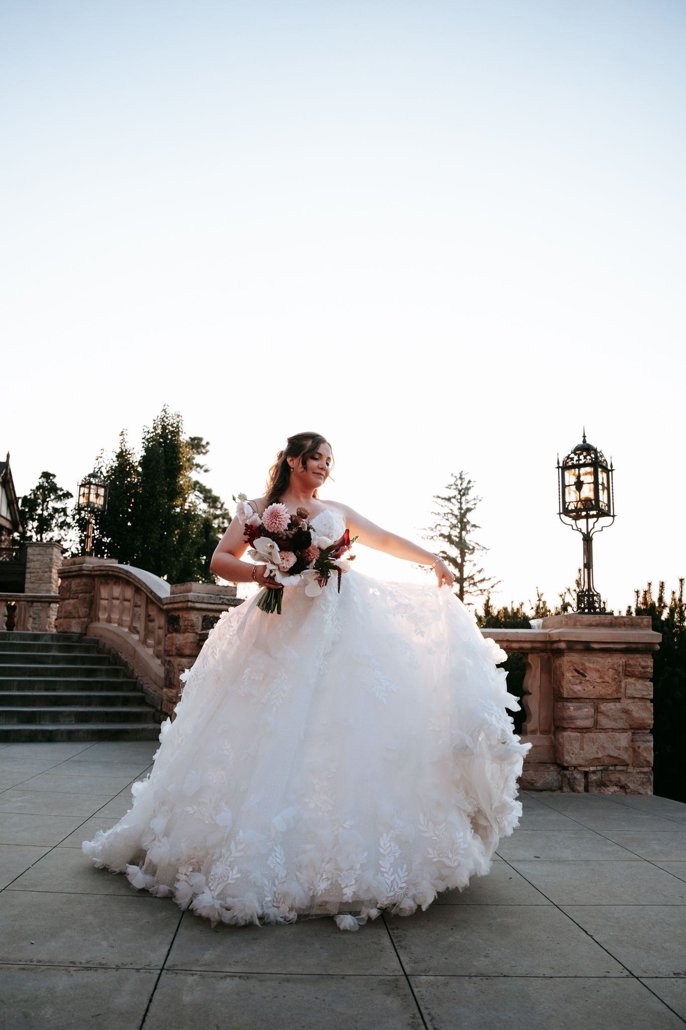 Bride twirling in a voluminous white ball gown while holding a bouquet on a stone terrace at dusk.
