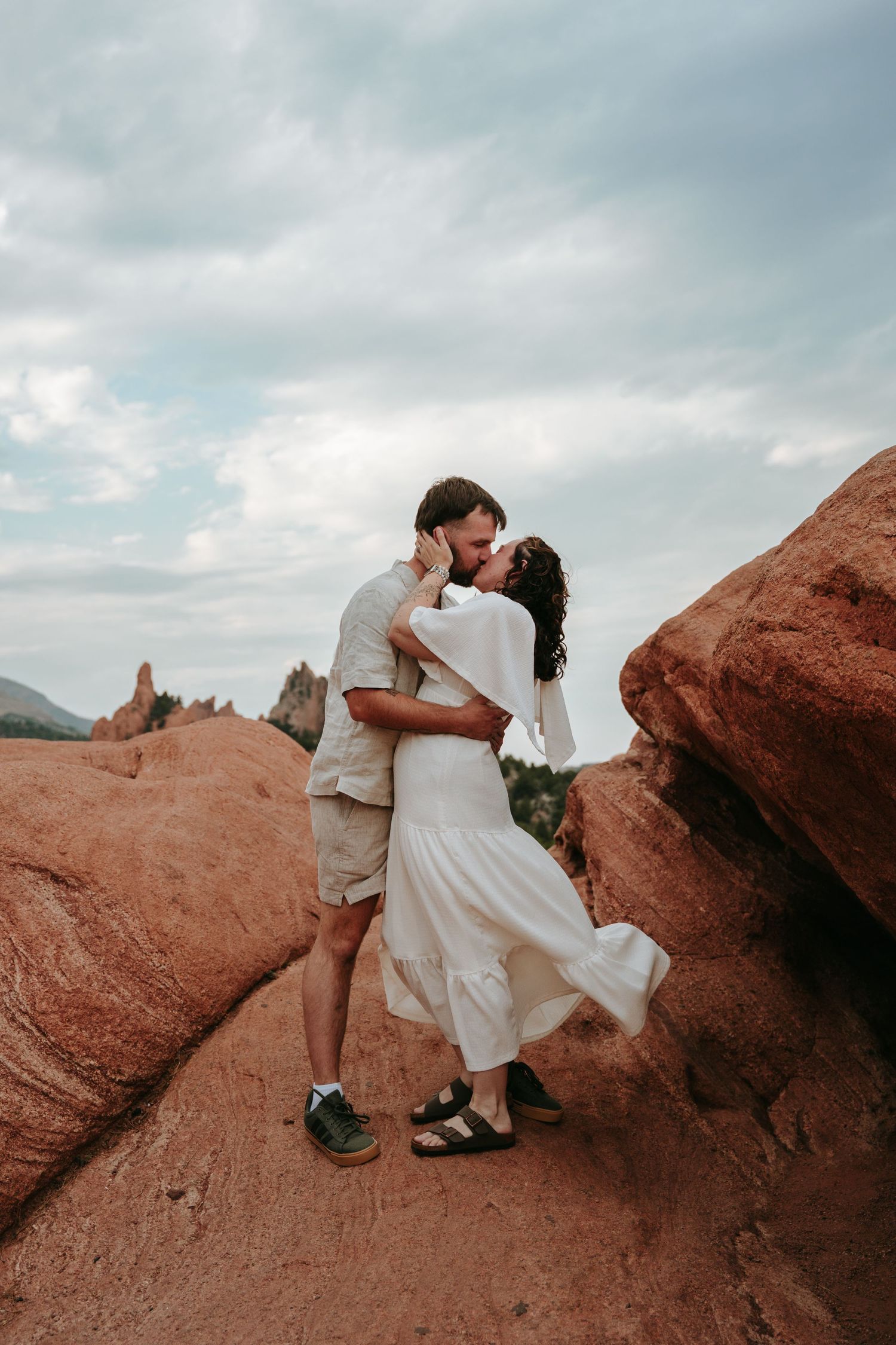 Couple kissing among red rock formations at Garden of the Gods, woman in white dress, cloudy sky backdrop.