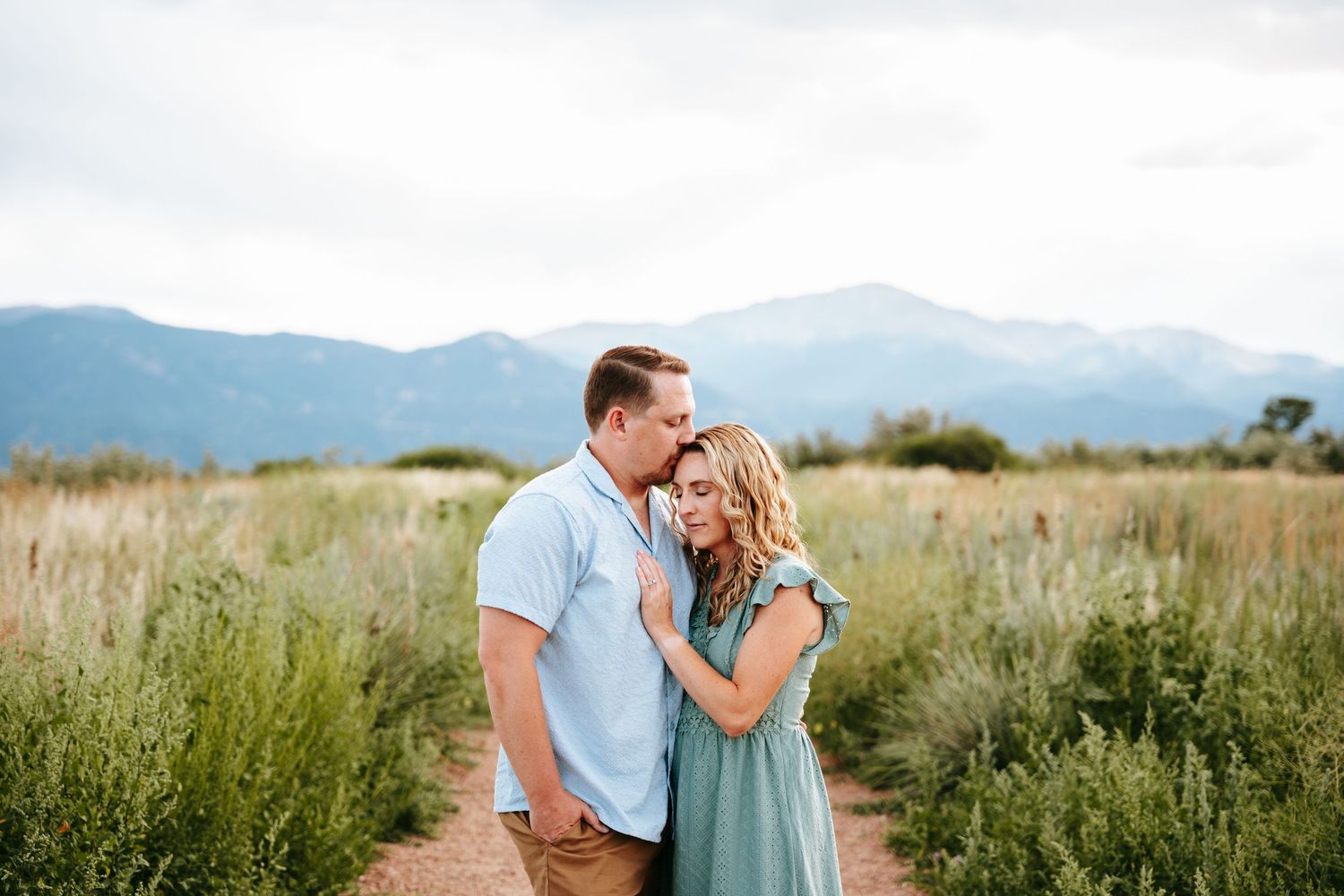 A couple sharing a tender moment on a dirt path surrounded by wildflowers and mountains.