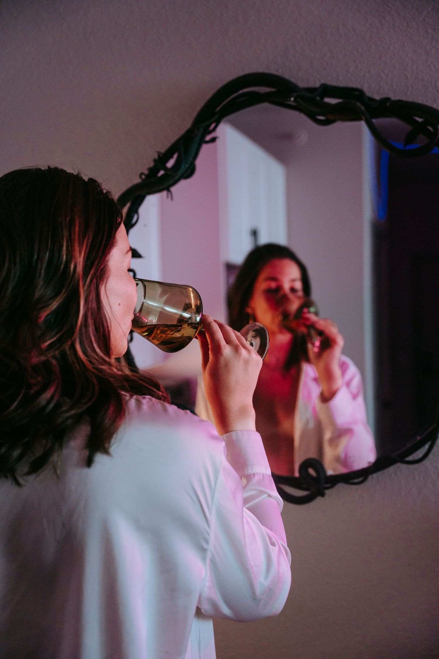 Woman in white robe drinking from glass wine goblet reflected in ornate mirror with purple lighting.