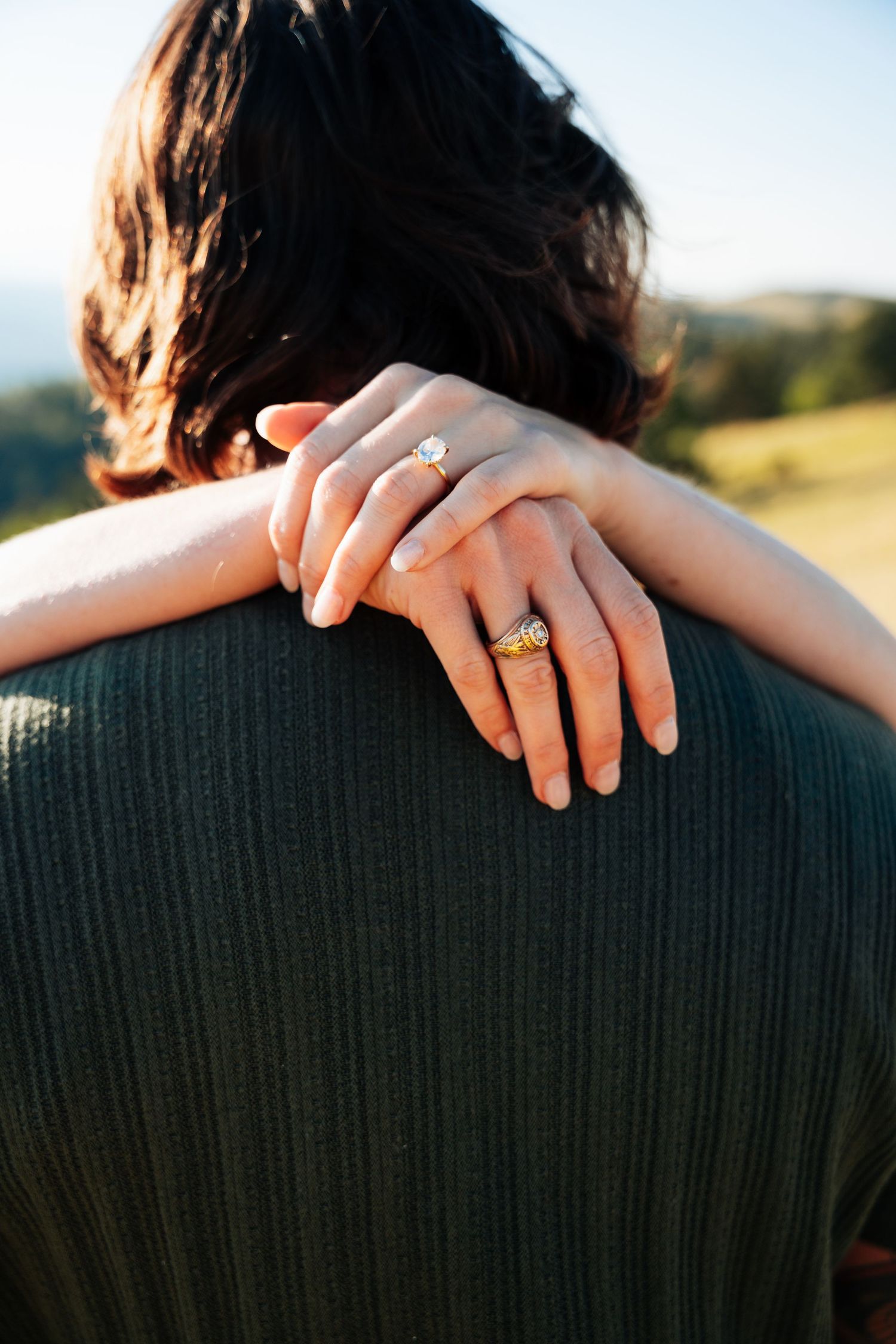 Close-up of woman's hands with oval diamond and gold band rings wrapped around her partner's green sweater.