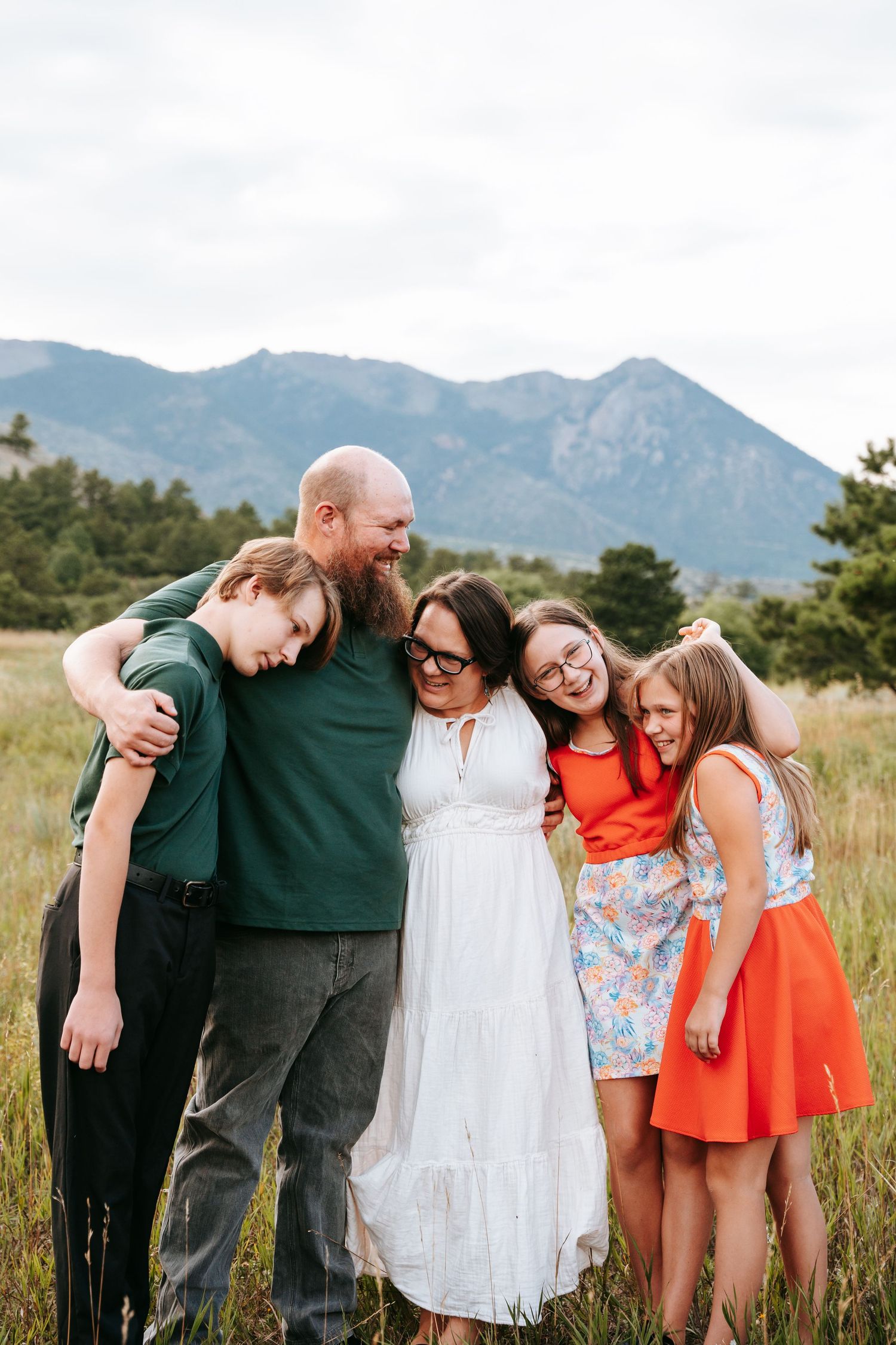 Joyful family of five embracing in mountain meadow, laughing together in colorful casual attire.