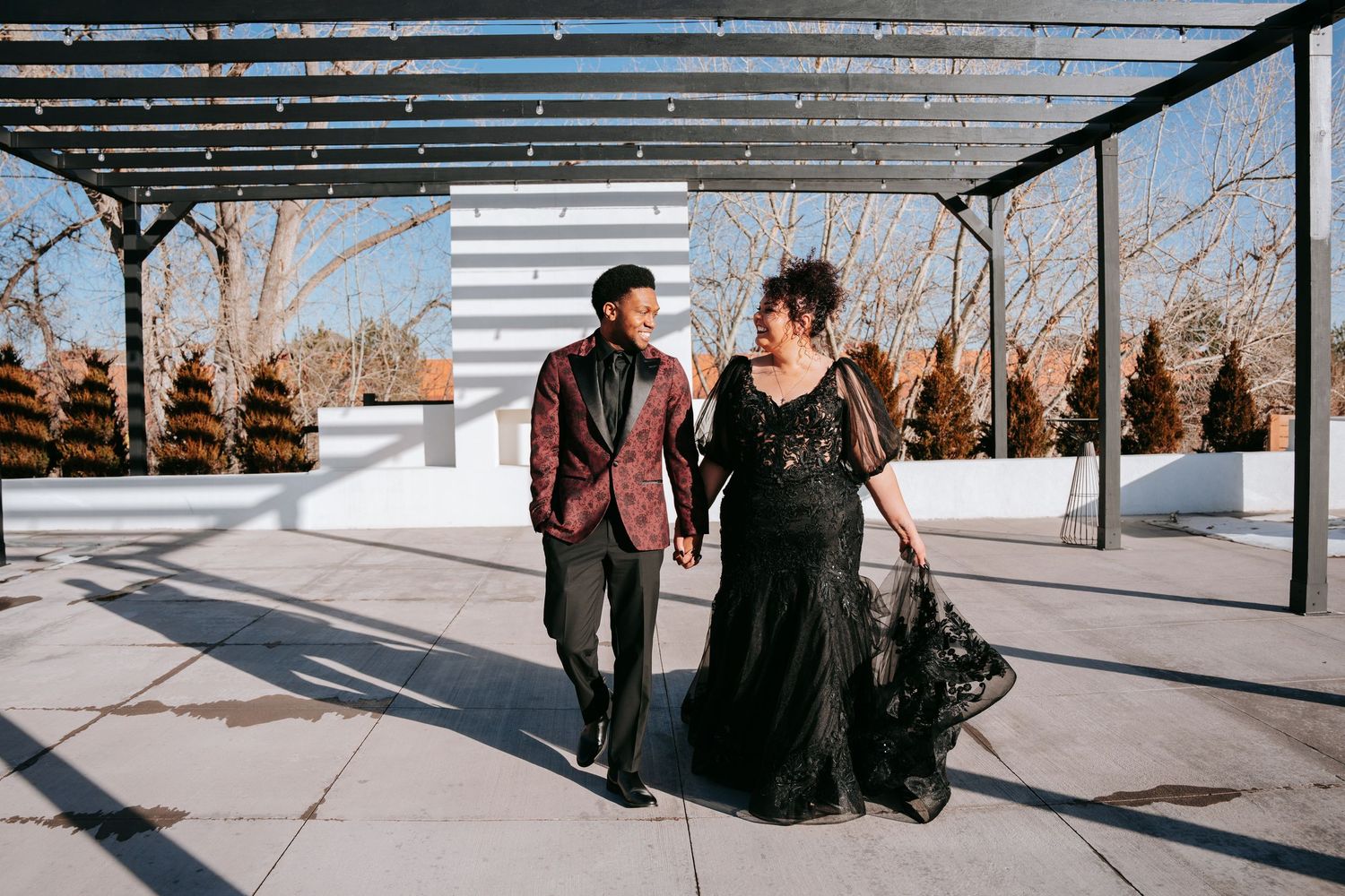 Couple in elegant black lace gown and red floral suit walking on rooftop terrace during winter engagement photo session.