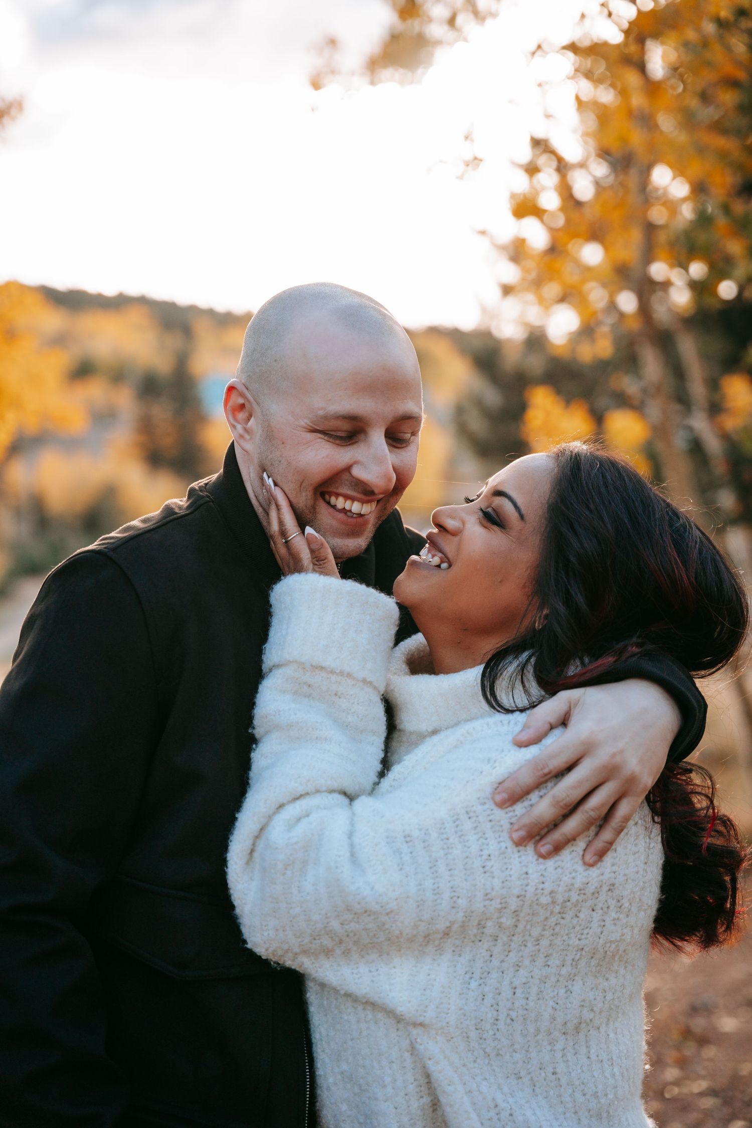 Couple laughing and embracing outdoors among golden autumn trees during a fall engagement photo session.