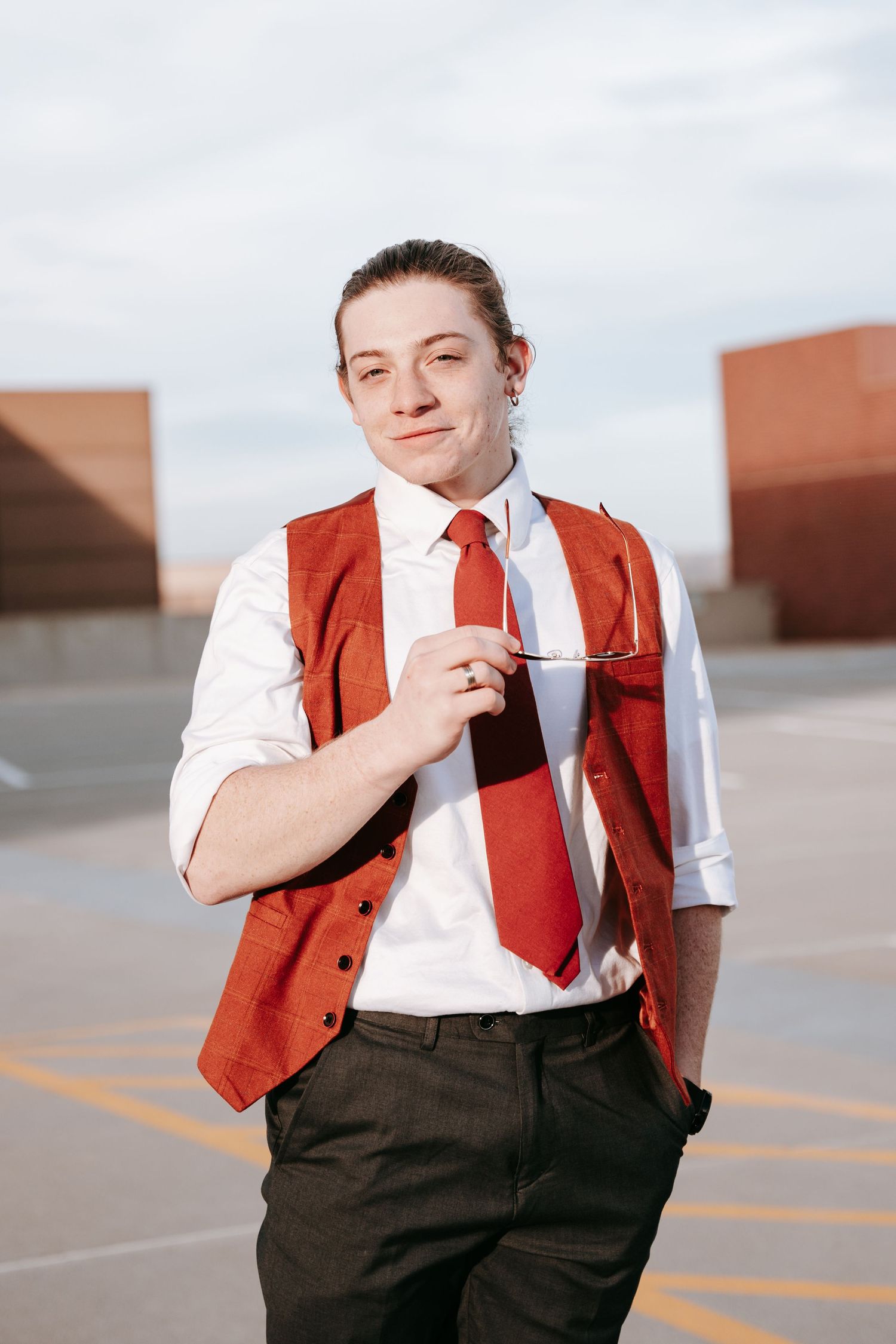 Young man in a red vest and red tie holding glasses on a rooftop parking lot.