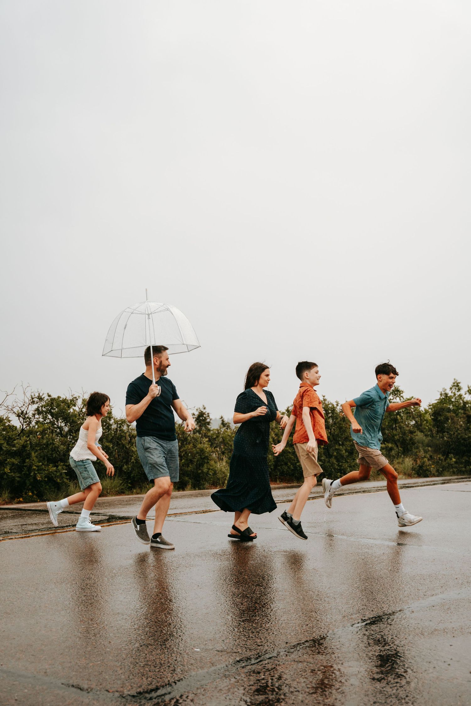 Family of five running across wet pavement in the rain, one adult holding a clear umbrella, trees in background.