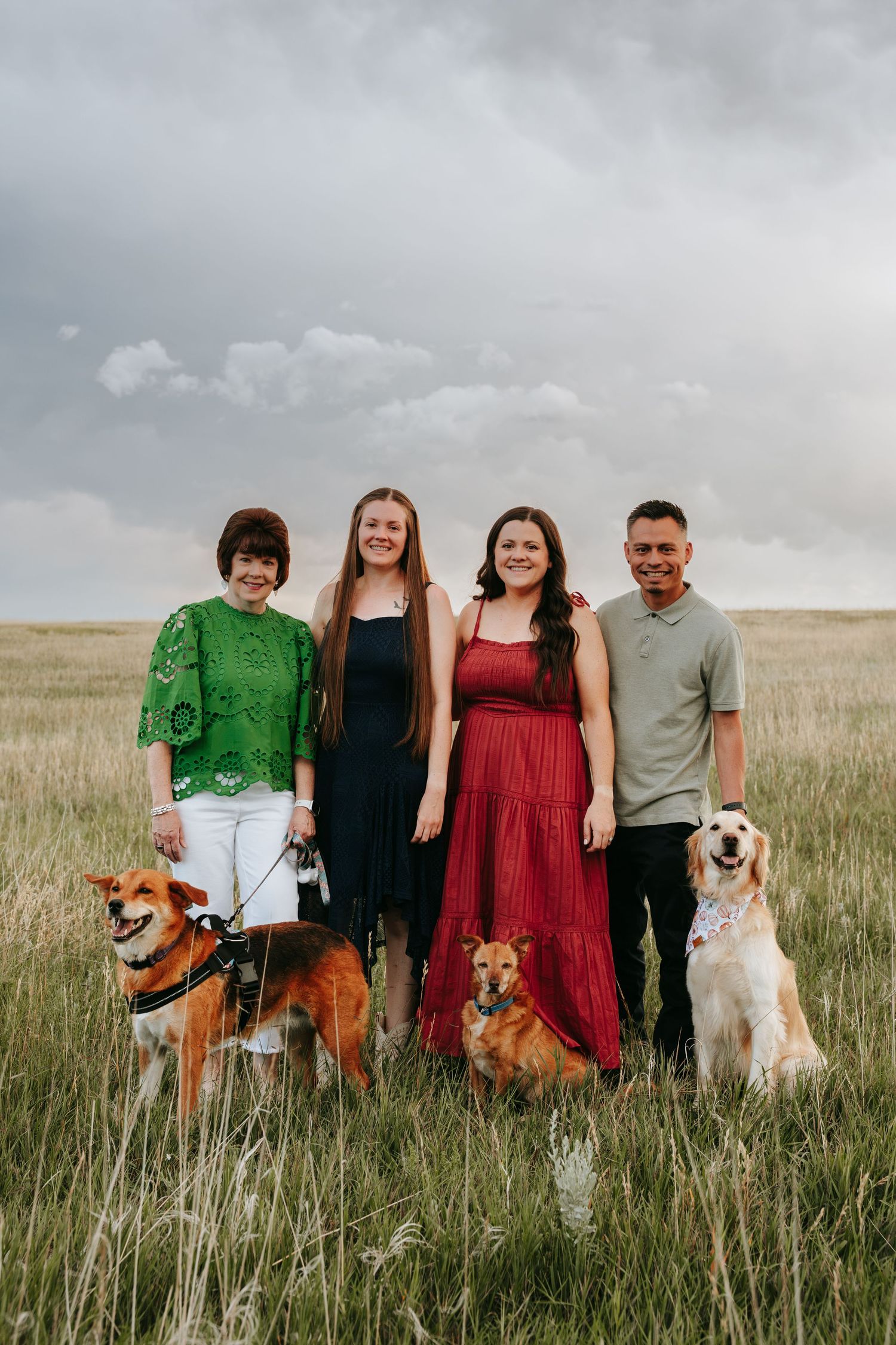 Family of four posing with three dogs in a grassy field under cloudy skies.