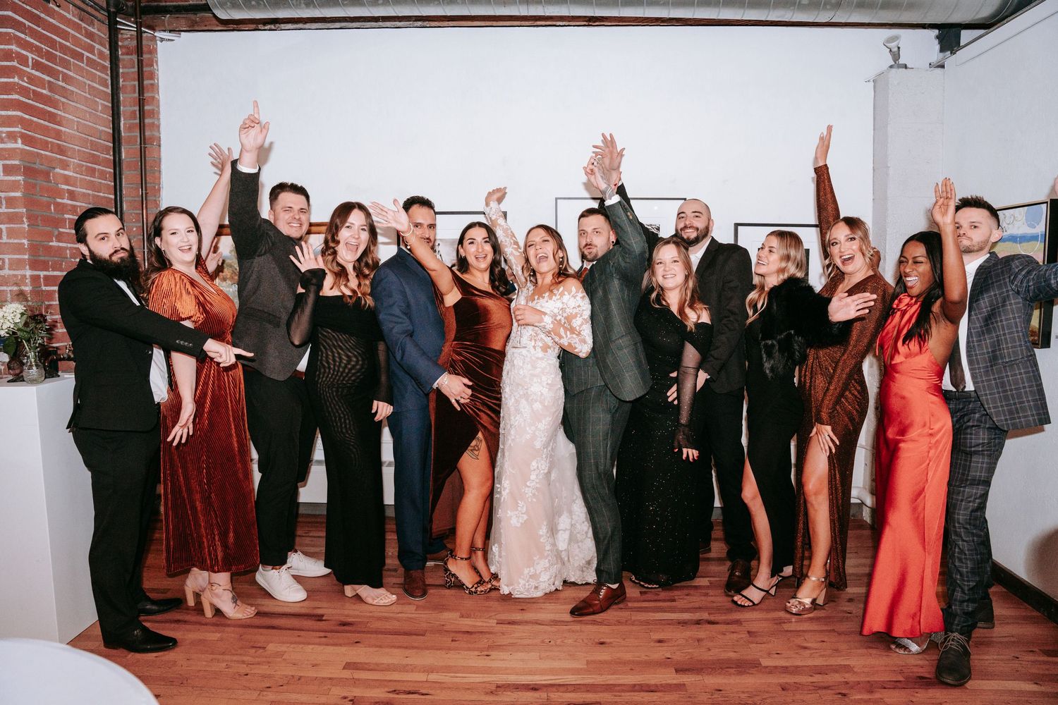 Bride and groom celebrate with their wedding party, all smiling with hands raised in joy.