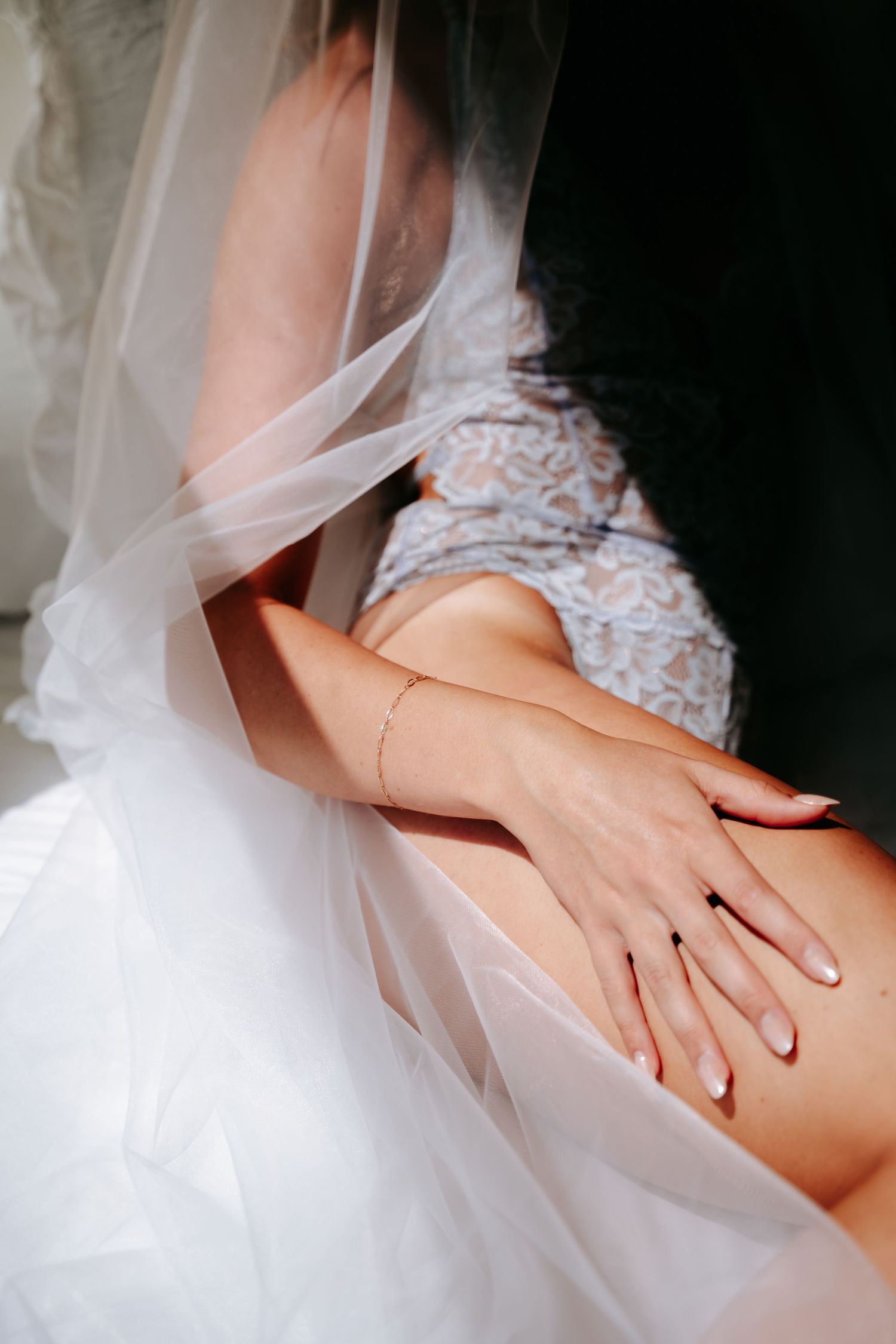 Bride in lace lingerie with wedding veil and bracelet, hands resting on lap, elegant bridal boudoir photo.