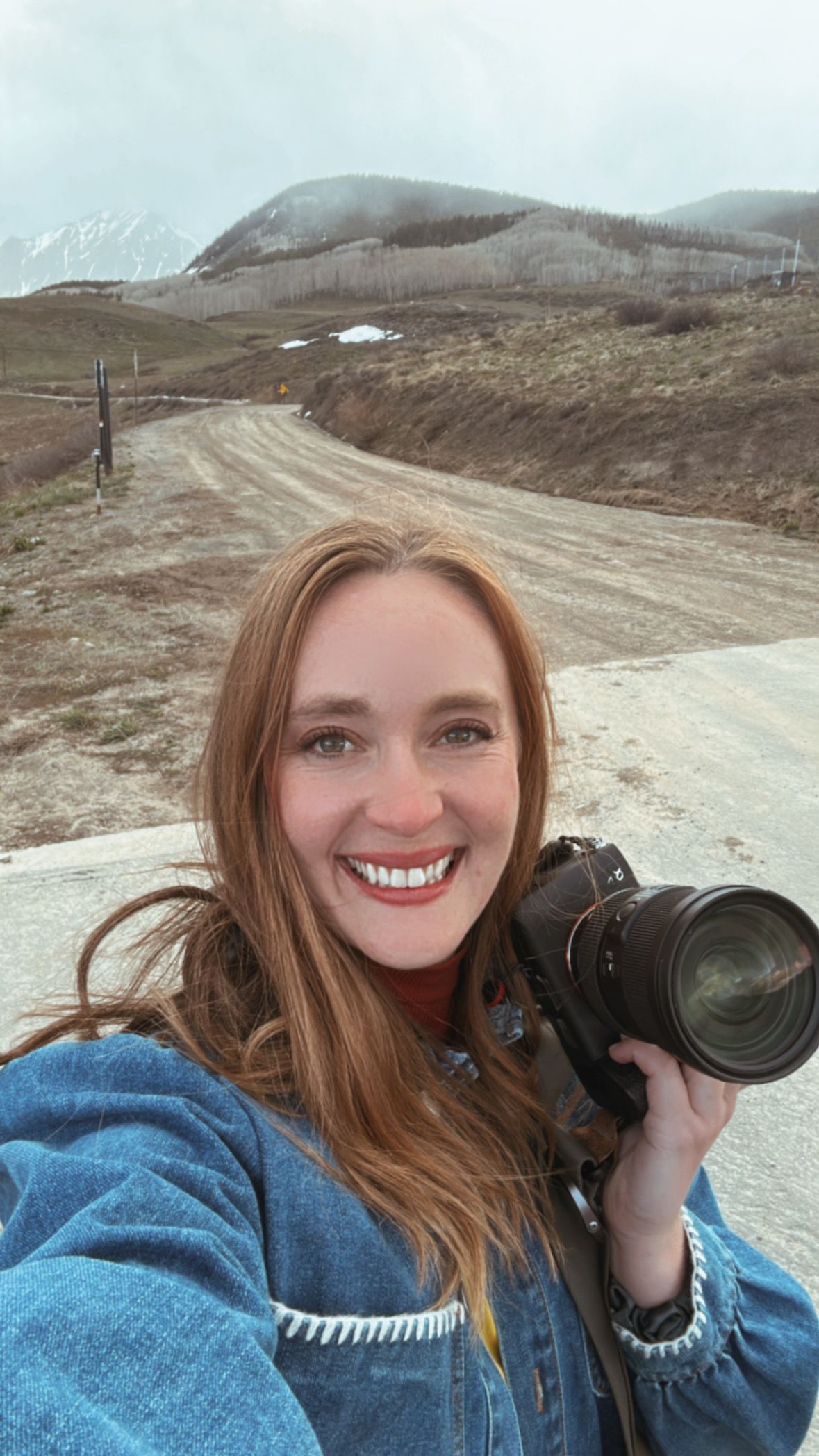 Smiling redhead female photographer holding camera taking selfie on mountain dirt road.