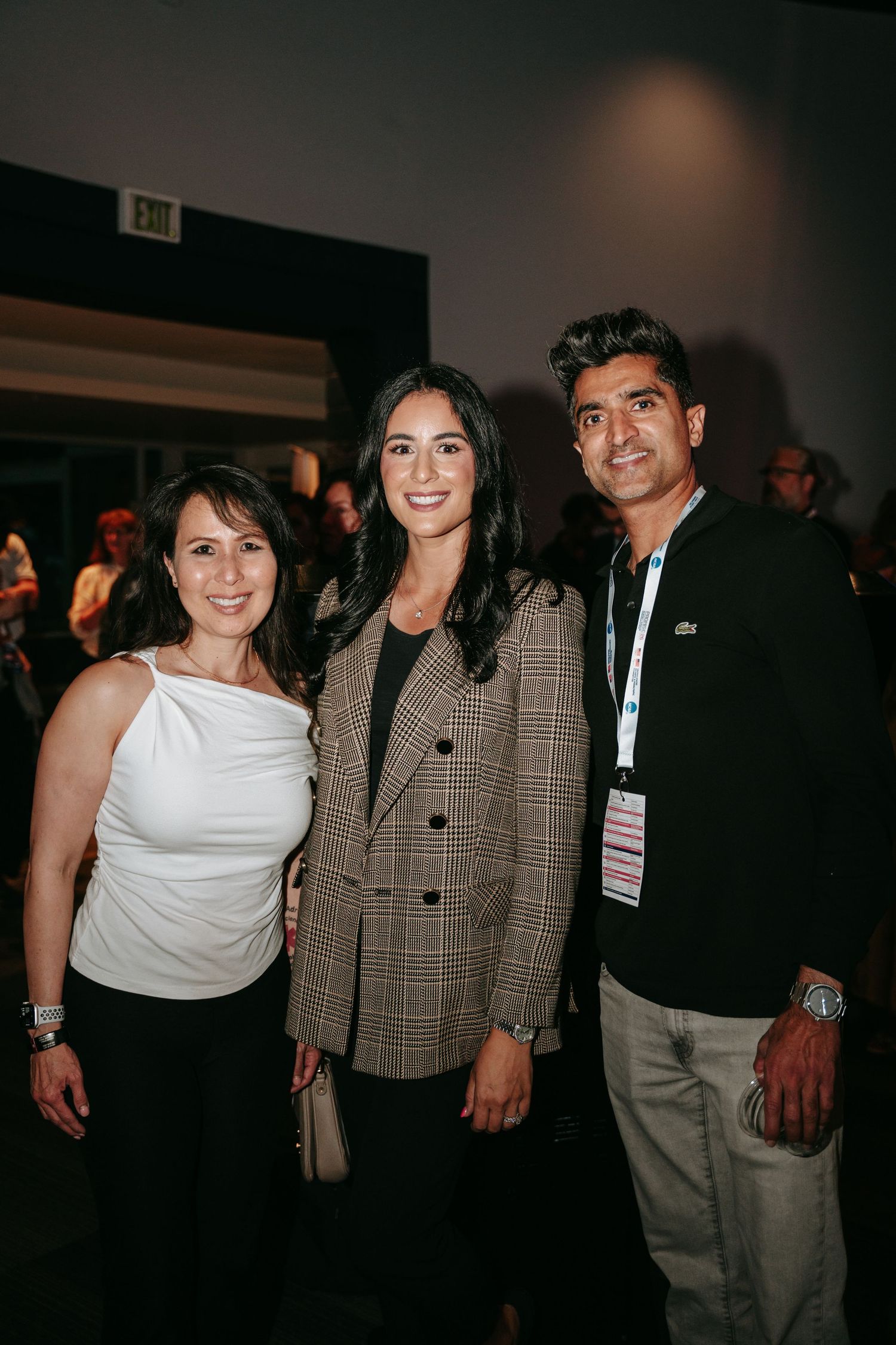Two women and a man pose smiling together at an indoor event, with the man wearing a conference lanyard and badge.