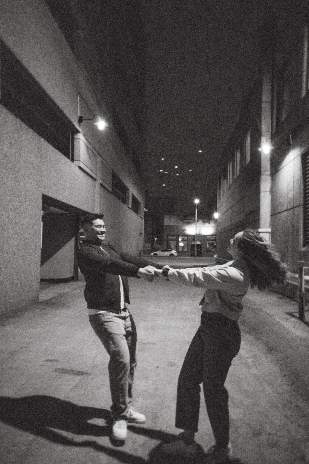Black and white photo of joyful couple spinning and dancing in illuminated city alley at night.