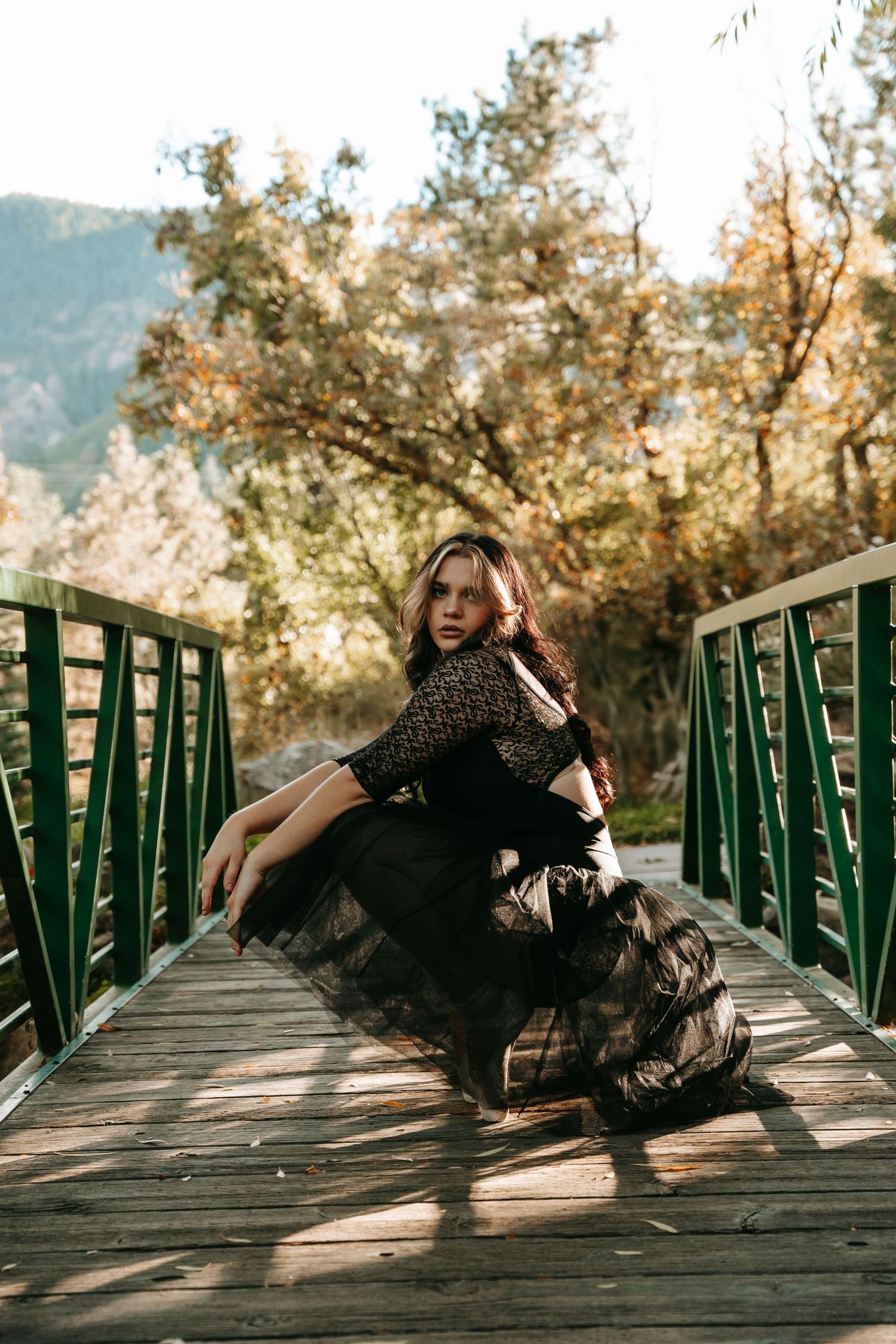 Woman in black tulle gown sitting on wooden bridge surrounded by autumn trees.