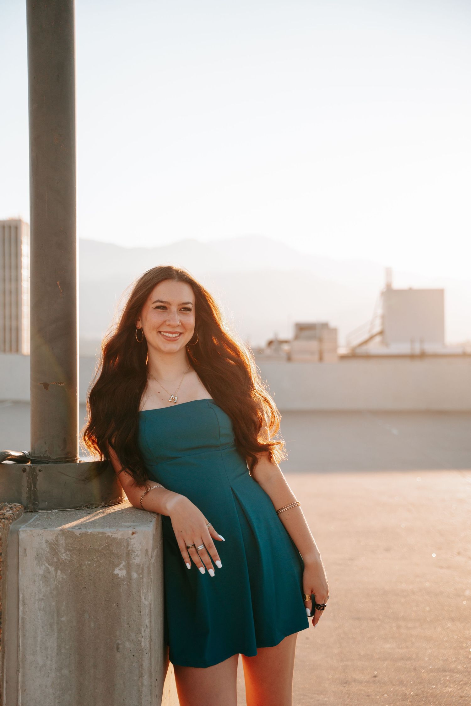 Smiling woman in teal strapless dress leaning against pillar at golden hour.