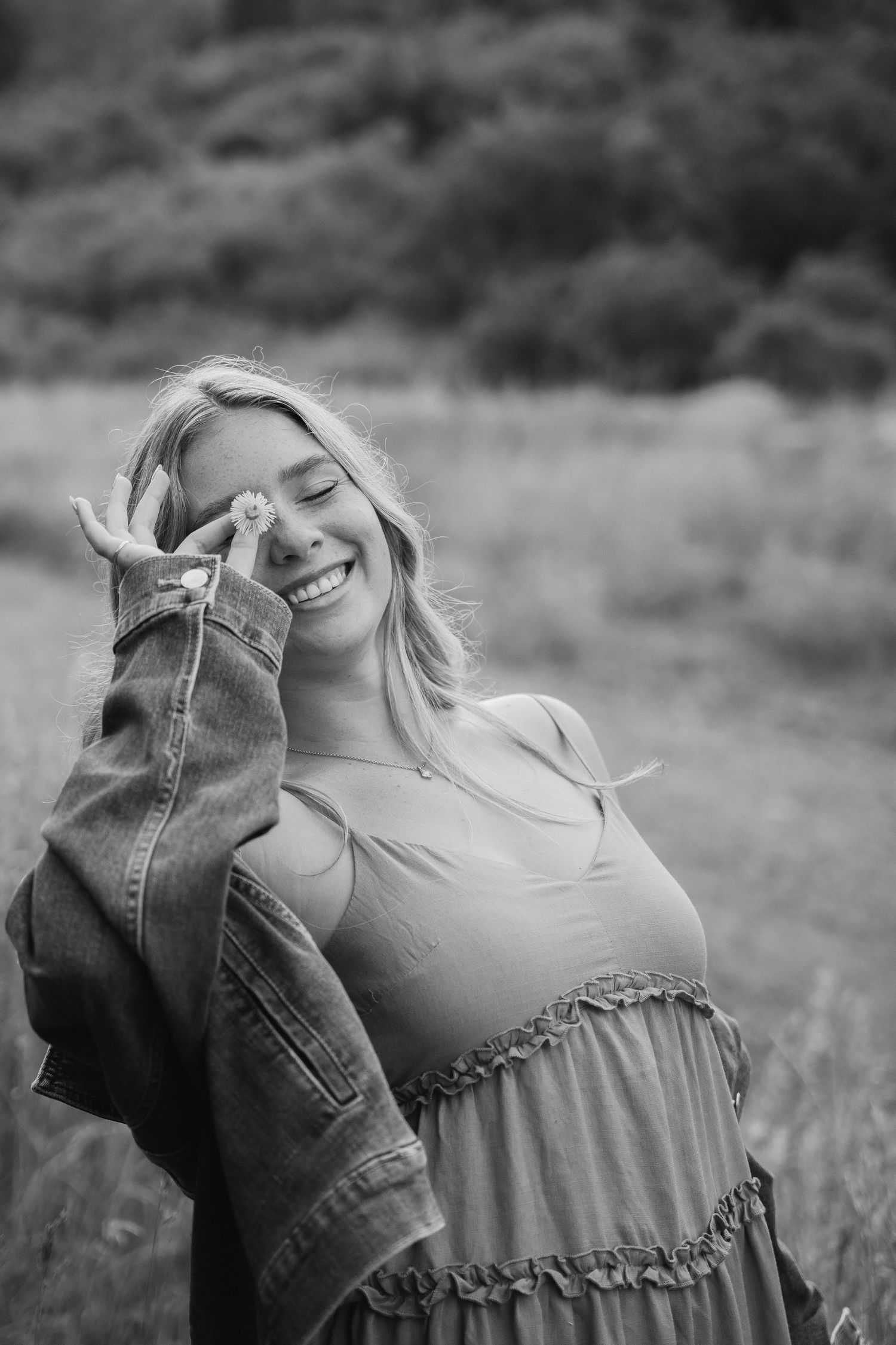 Black and white photo of a smiling blonde woman holding a flower to her face outdoors.