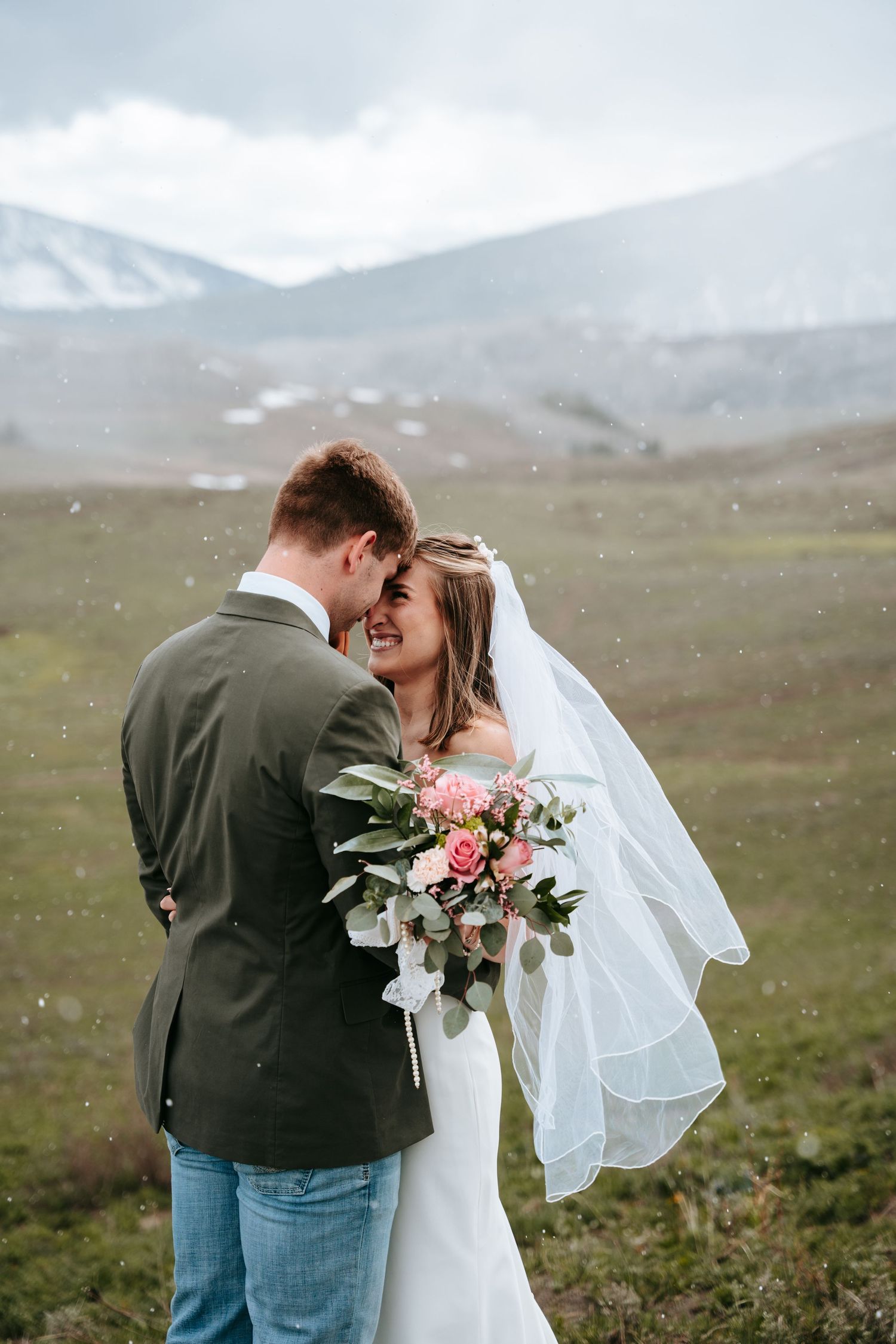 Bride and groom sharing romantic moment in snowy mountain landscape, bride holding pink floral bouquet.