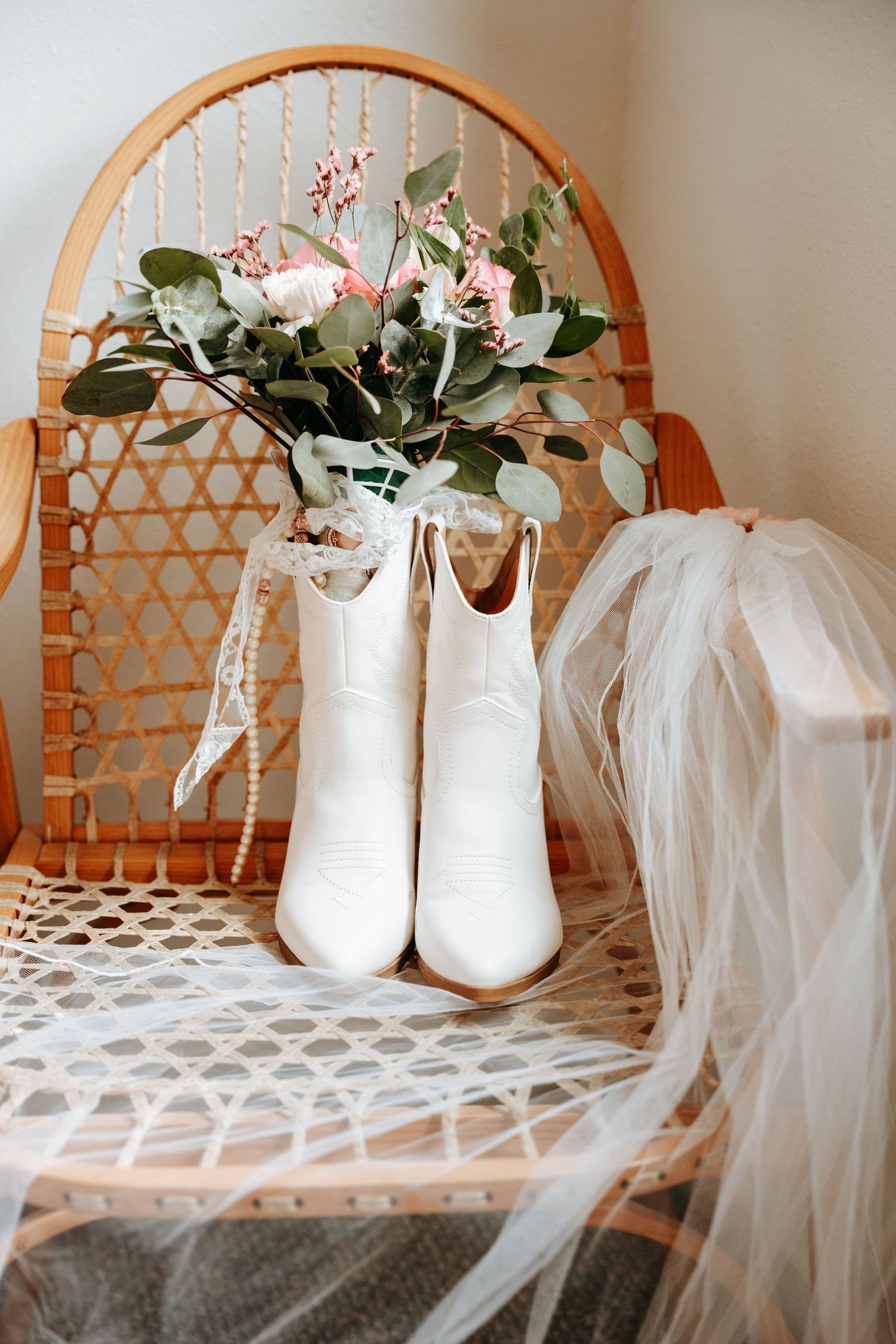 White bridal cowboy boots with pink floral bouquet and veil displayed on wicker chair.