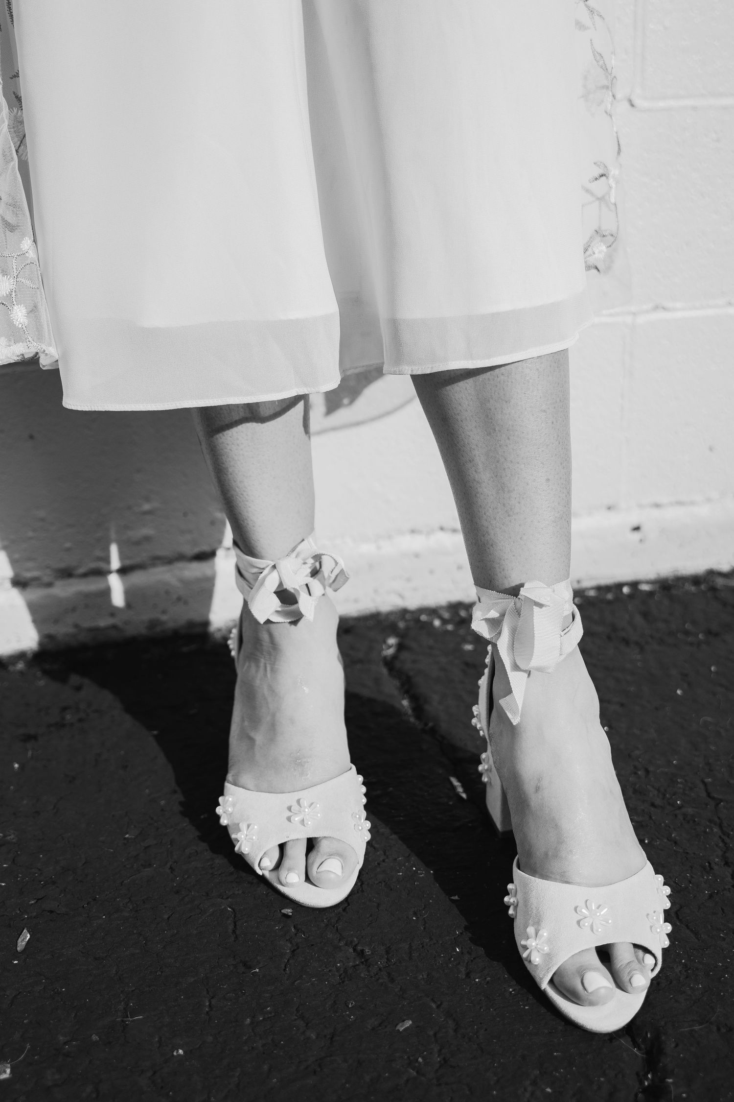 Black and white close-up of bride wearing floral pearl-embellished heeled sandals with ribbon ties.