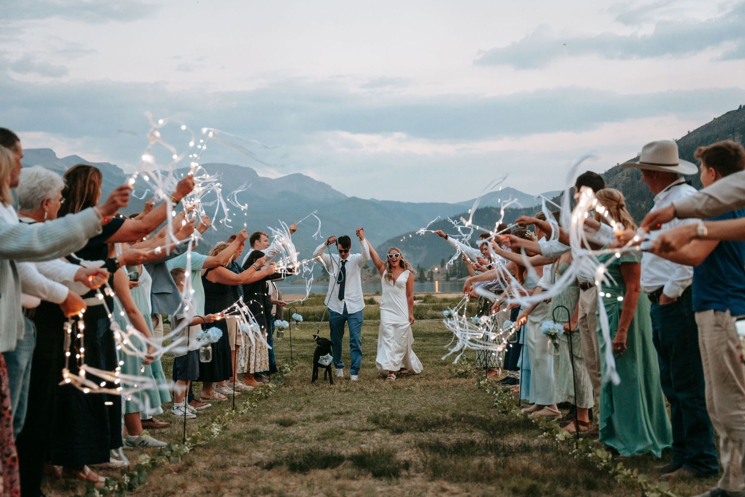 Bride and groom walk through ribbon wand sendoff with mountain backdrop at outdoor wedding ceremony.