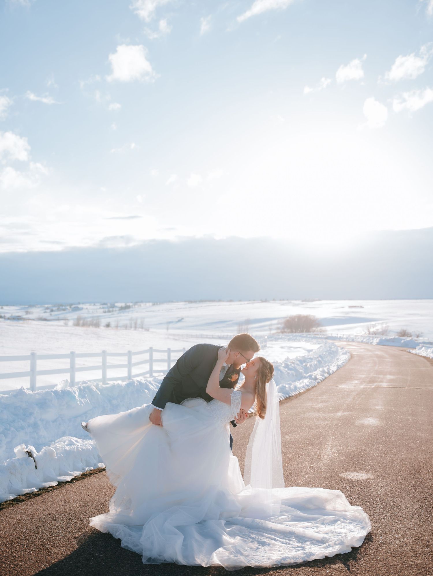 Bride and groom sharing a dip kiss on sun-drenched snowy road with dramatic winter sky backdrop.