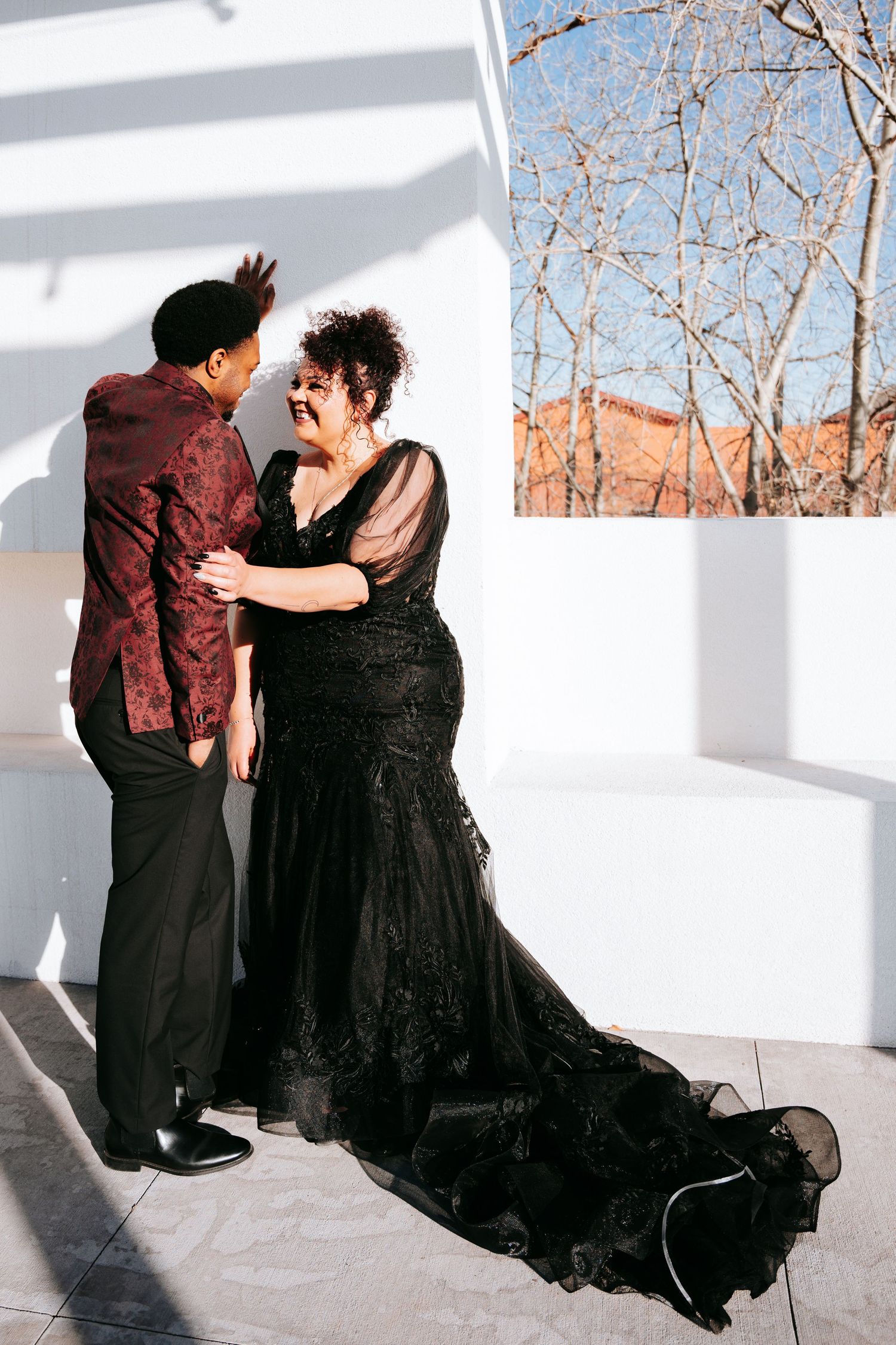 Bride in elegant black lace mermaid gown with train dancing with groom in burgundy floral shirt outdoors.