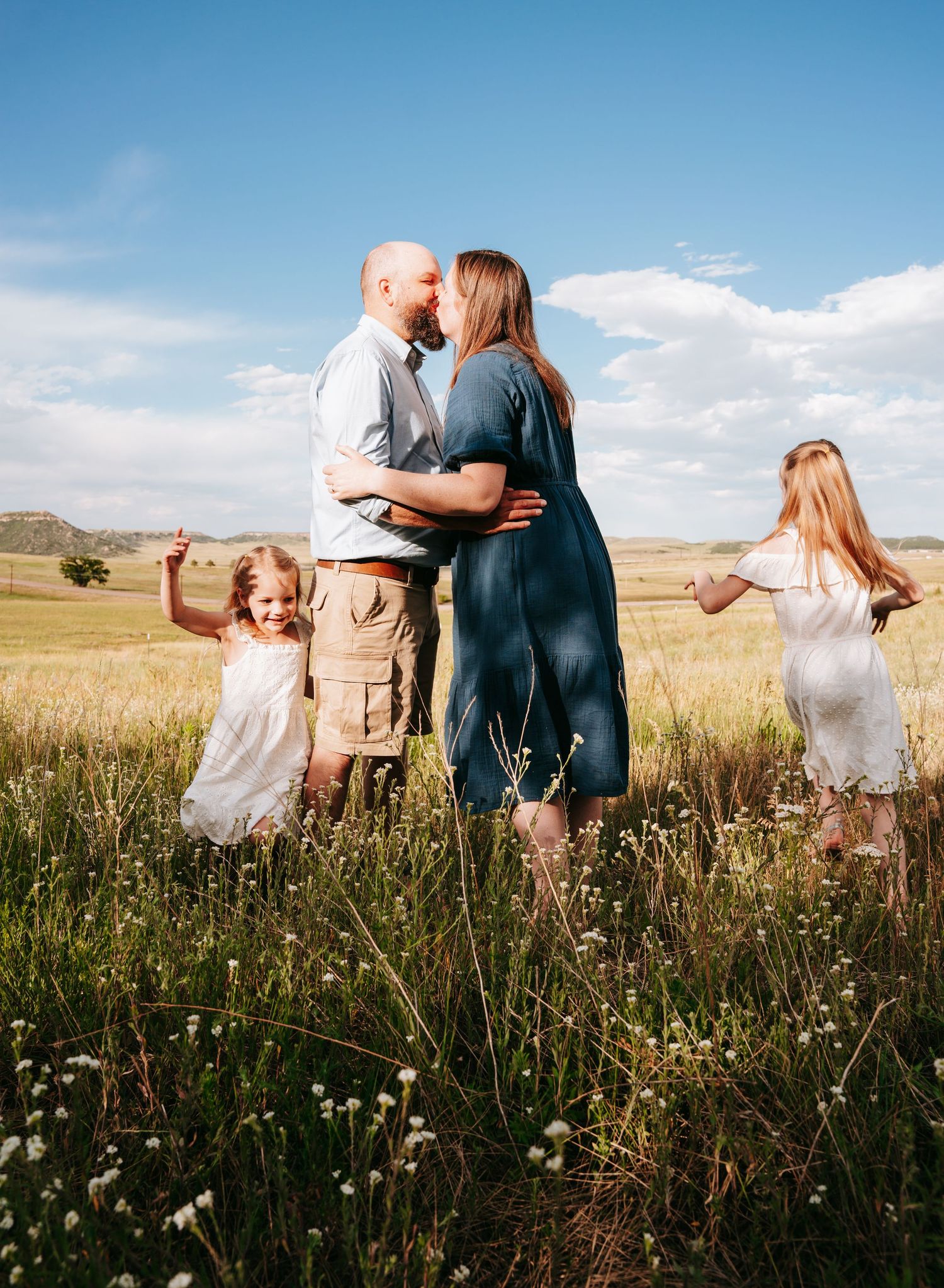 Couple sharing a kiss in sunny meadow while two young daughters in white dresses play around them.