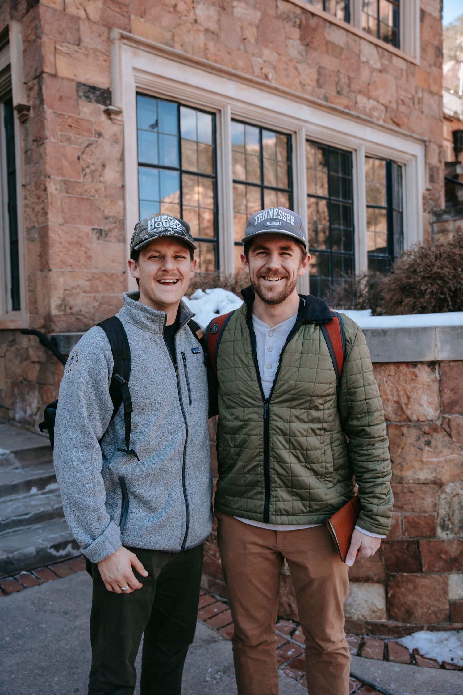 Two smiling men in baseball caps and jackets with backpacks stand outside a stone building with large windows on a snowy day.