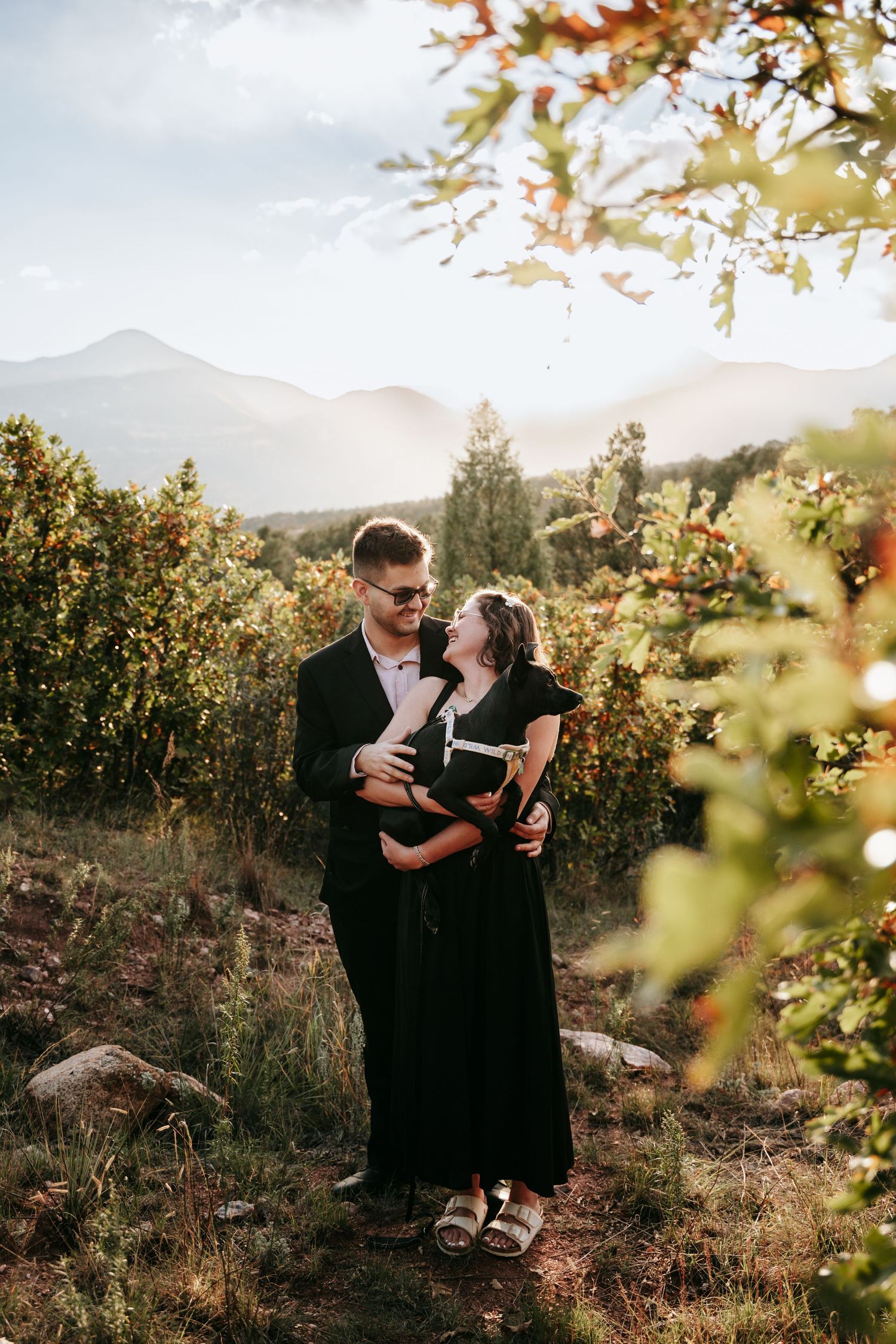 Couple in black outfits embracing and smiling at each other on a sunlit hillside with autumn foliage and mountains in the background.