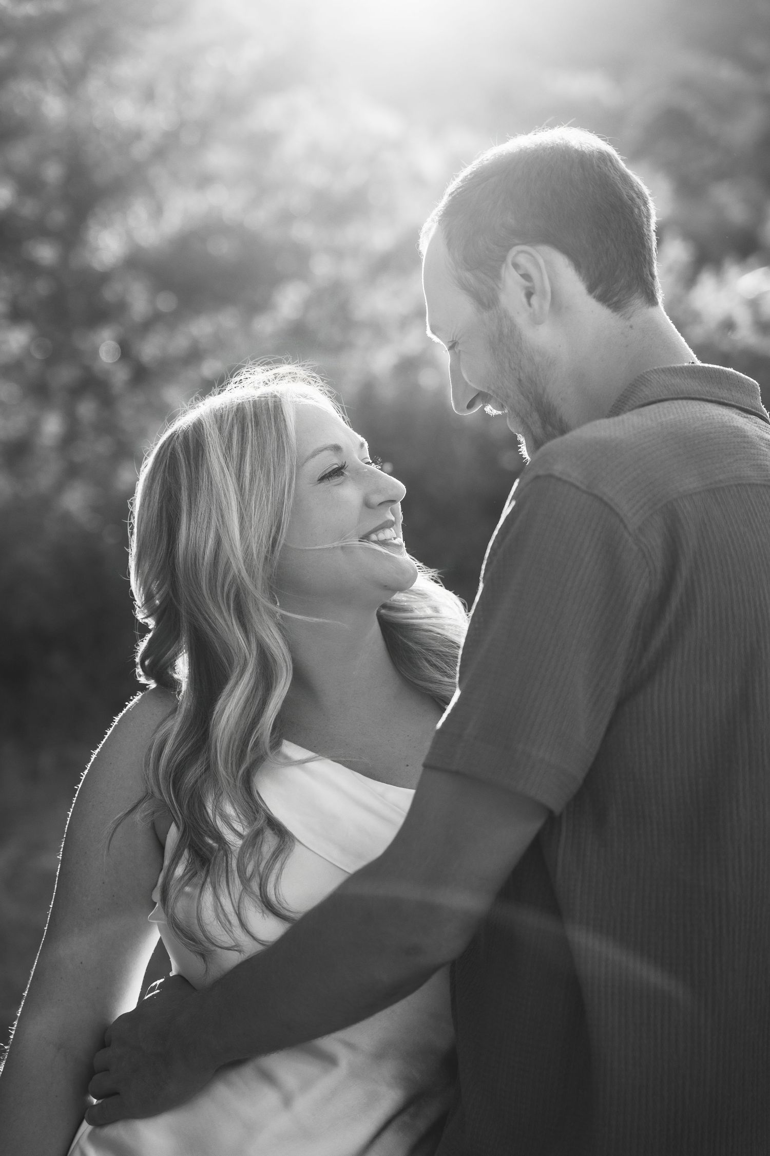 Black and white photo of a smiling couple embracing outdoors with sunlight glowing behind them.