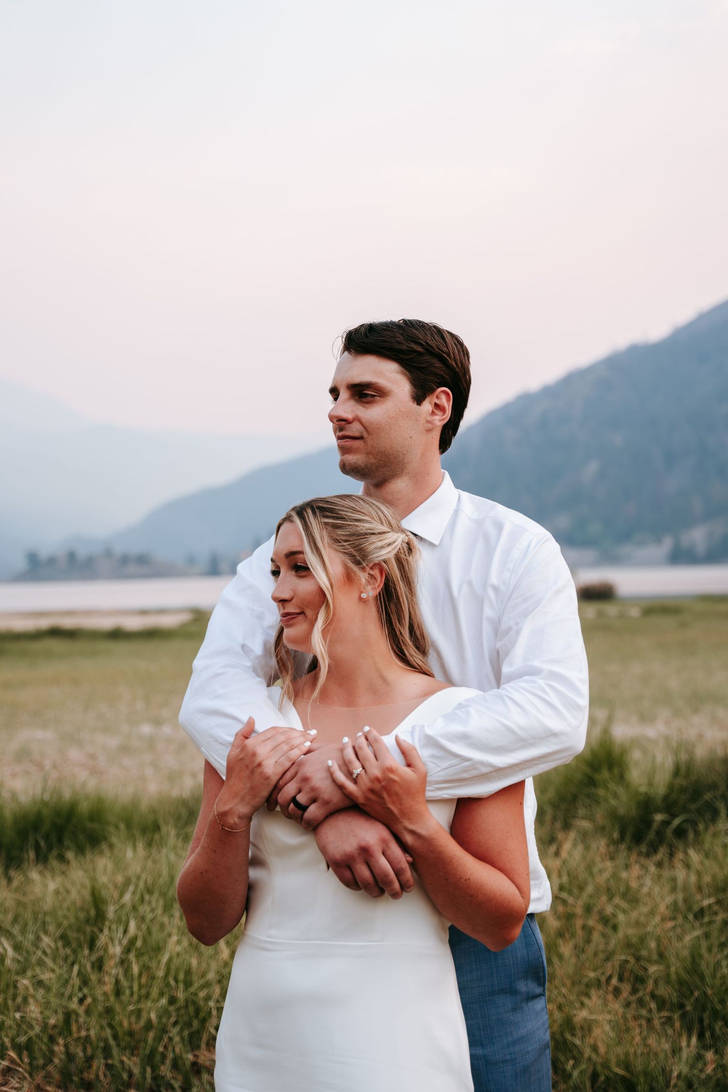Groom in white shirt embraces bride in white dress while gazing at mountain lake at dusk.