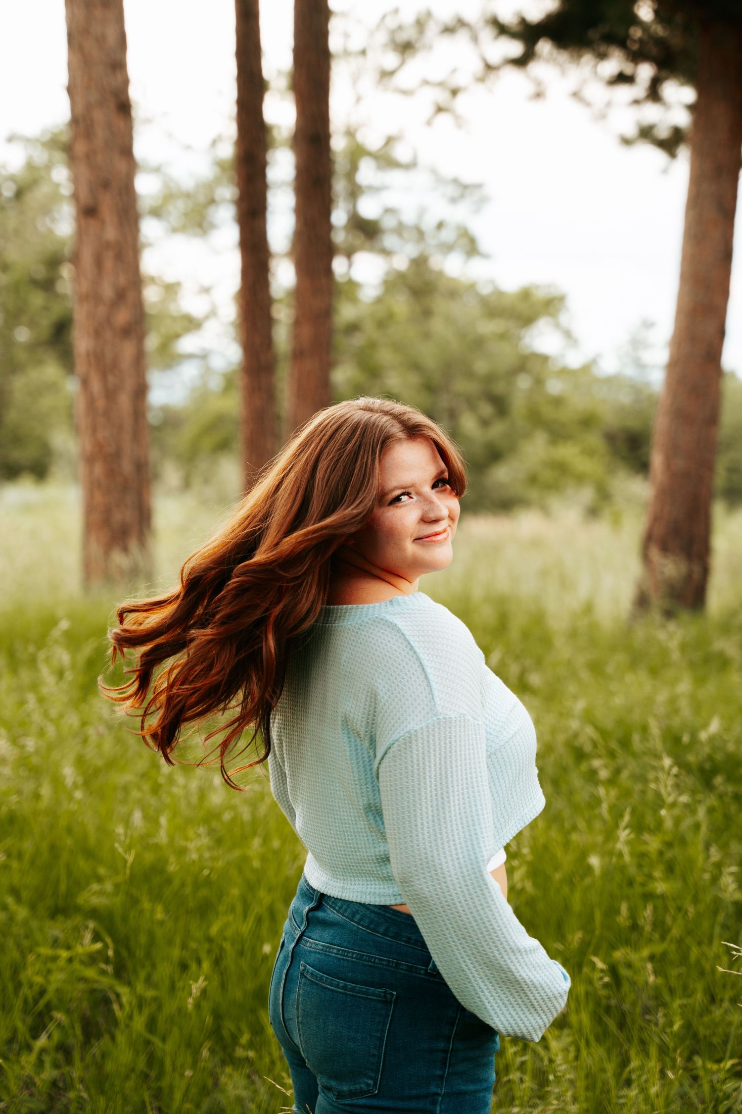 Young woman with flowing auburn hair in mint top and jeans smiling in a lush green forest setting.