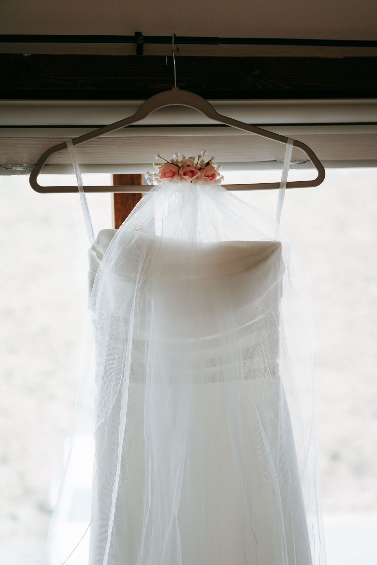 White strapless wedding dress with veil and pink floral accent hanging on hanger by window.