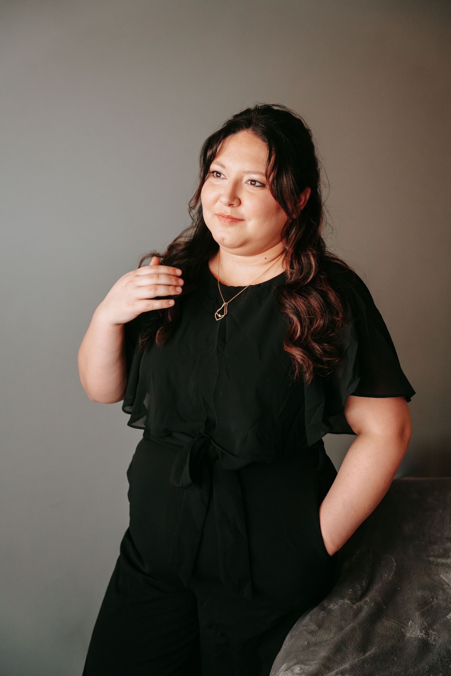 Young woman in black jumpsuit posing confidently against a gray background with gold necklace.